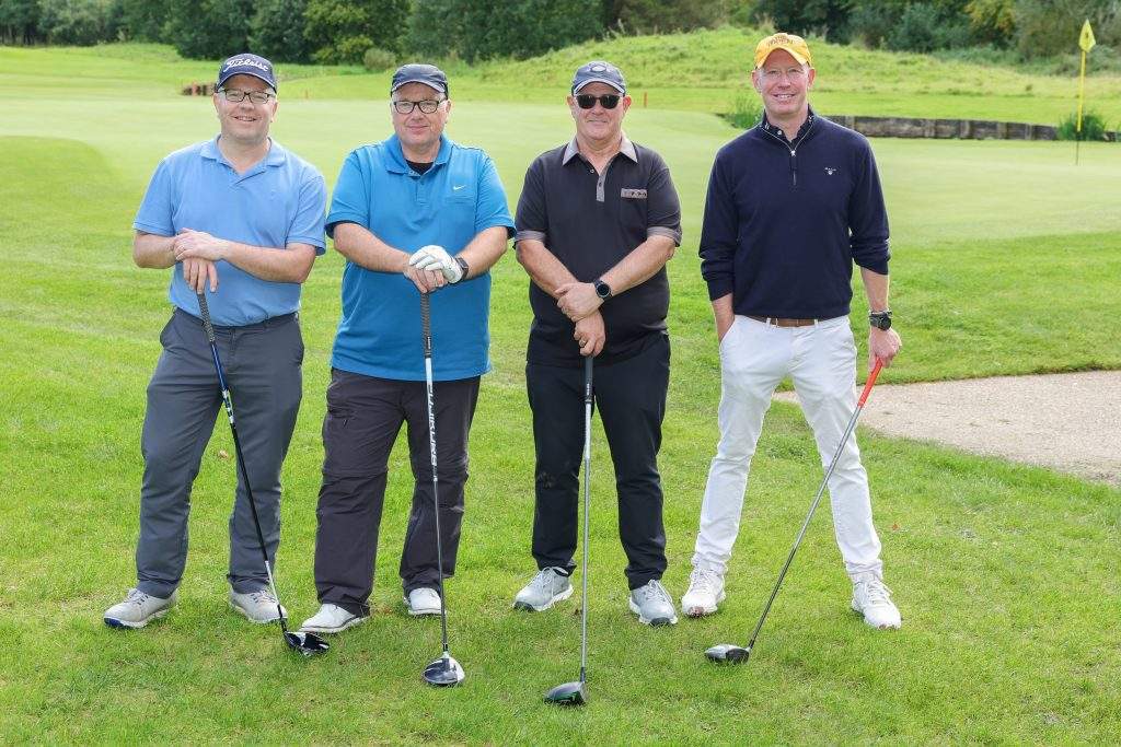 Four men standing on the green in a line with their golf clubs. 