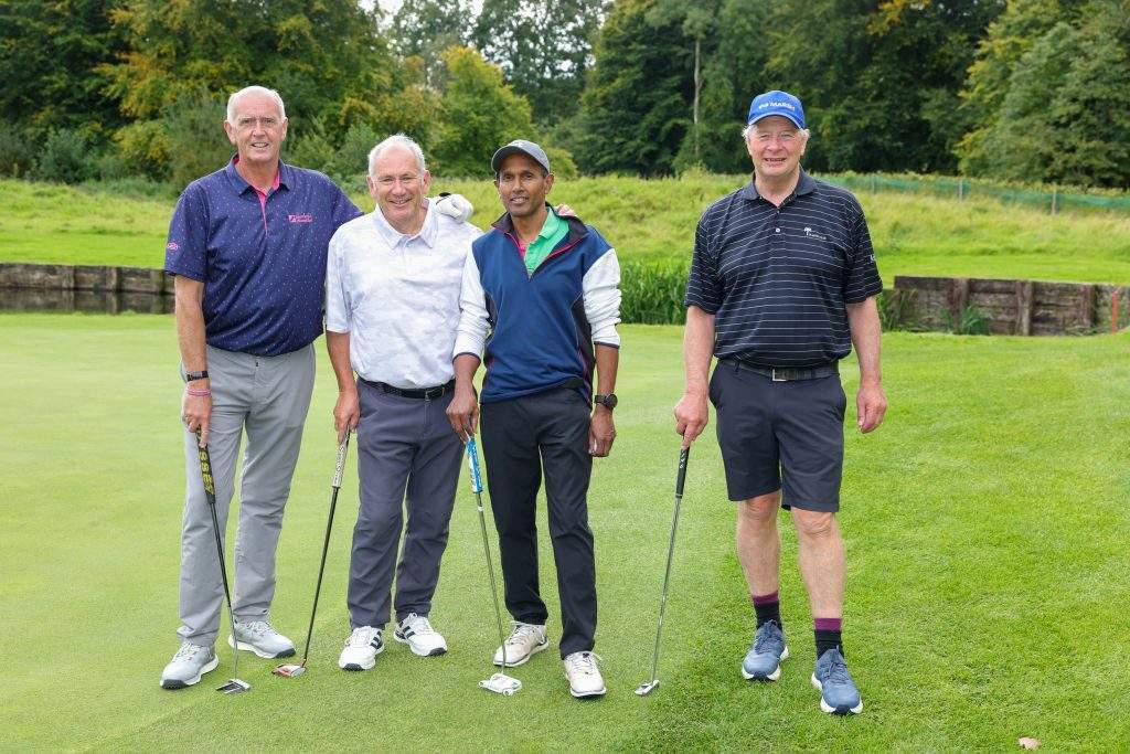 Four men standing on the green in a line with their golf clubs, arms around their shoulders with the water in the background.