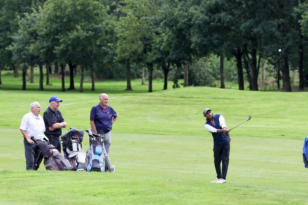 Four golfers on the green. Three watching one golfer take a swing.