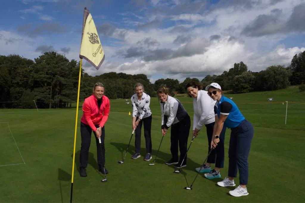 Five female golfers posing on the green with golf clubs; Galgorm flag at the front with NI Chamber's CEO, Suzanne Wylie amongst the golfers.
