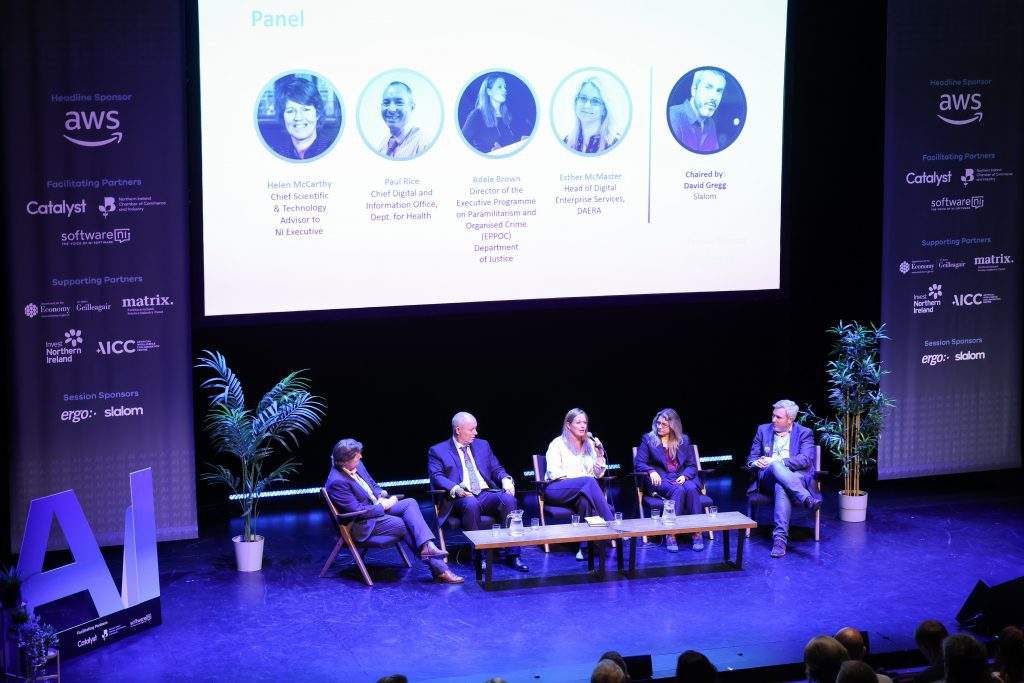full panel speaking on stage with a screen above them showing their profiles