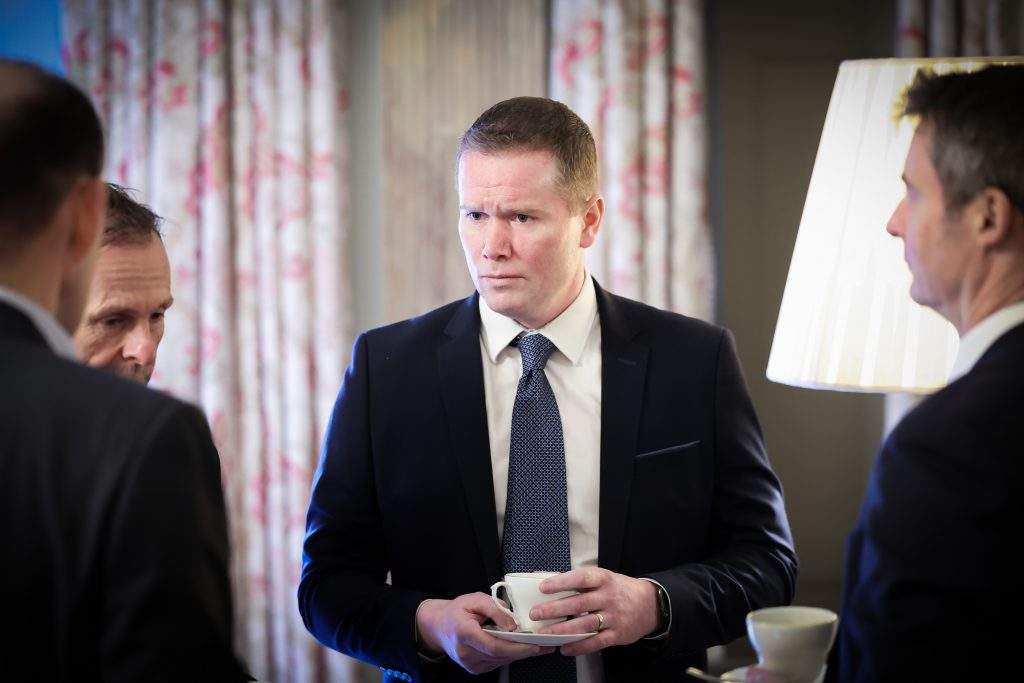 A person in formal clothing holding a cup and saucer while speaking with others in a room with patterned curtains.