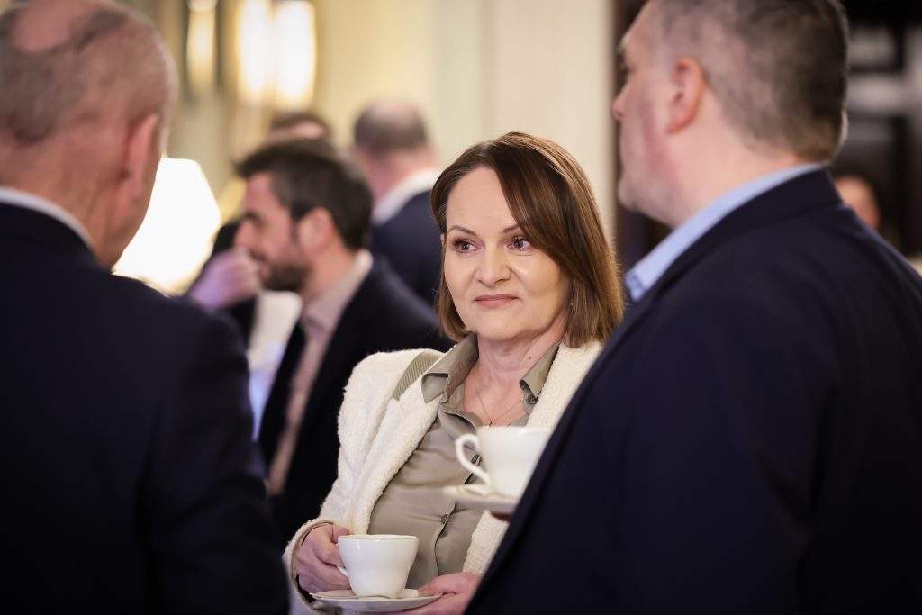  People standing together indoors at a networking event, holding cups and saucers while talking.