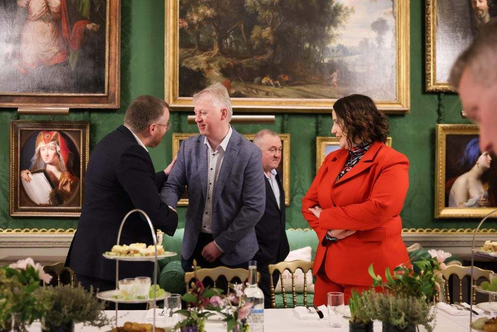  A person shaking hands with another attendee at a dining table, with framed artwork on the walls and table decorations visible.