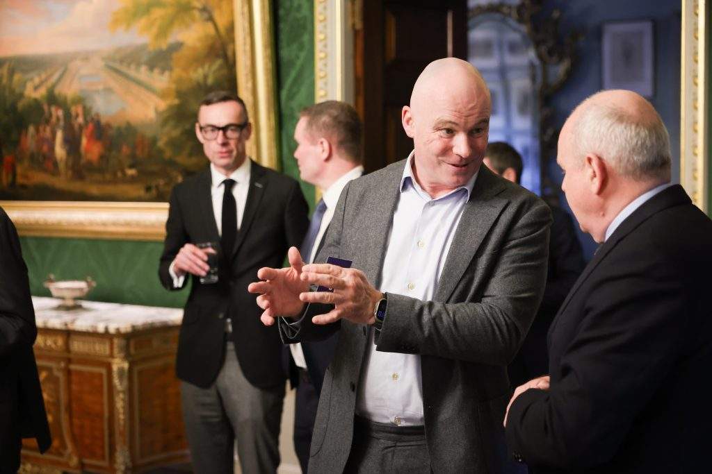 Several people in conversation during a formal event, with one person gesturing while others listen, surrounded by ornate decor and a large wall painting.