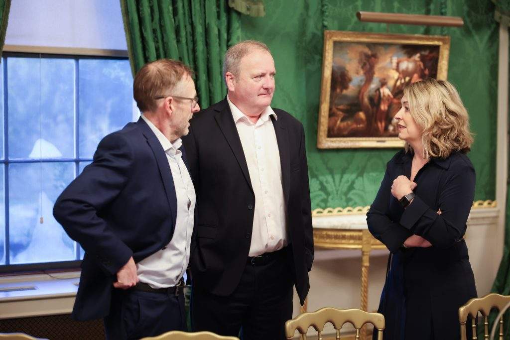 Three people standing together and talking beside a window in a formal room with green drapes and framed artwork.