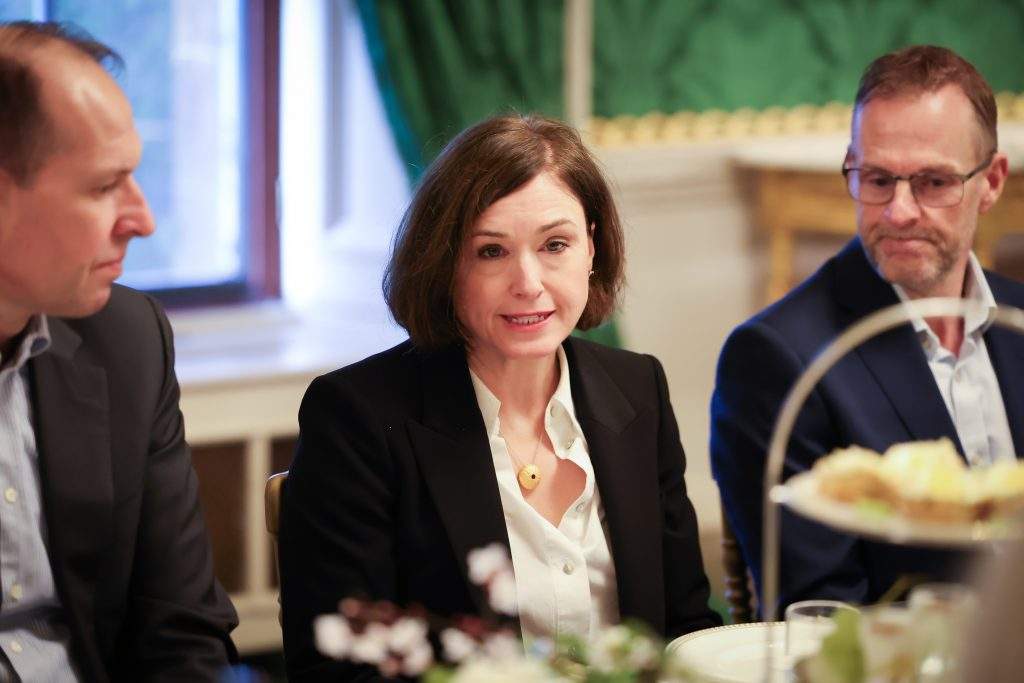 A group of people seated at a dining table, with one person sitting near a window with green drapes.