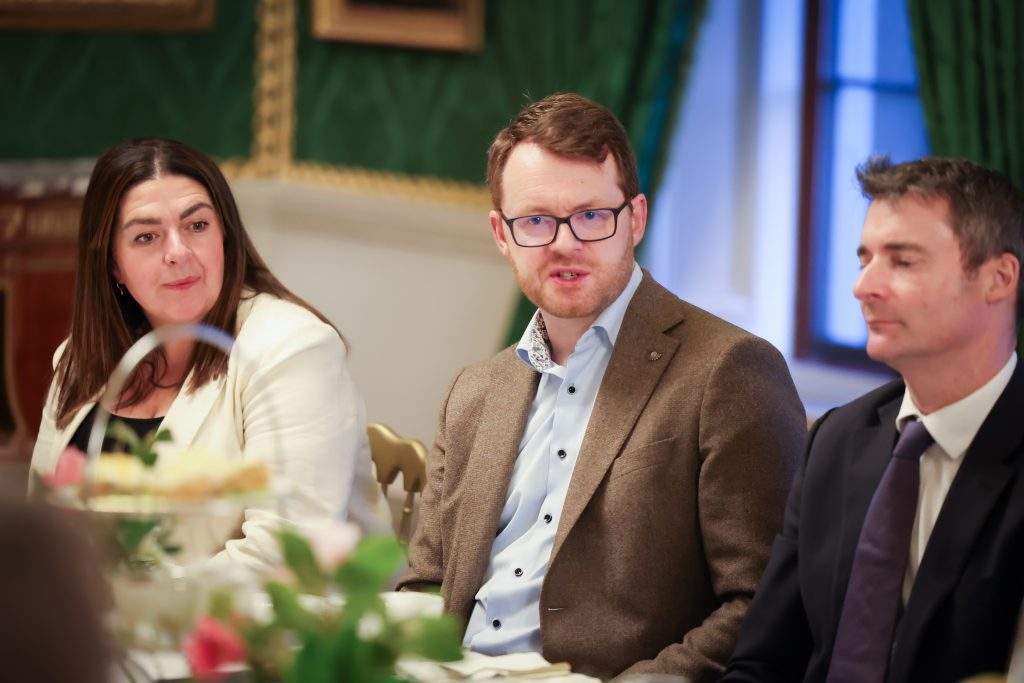 Three people seated at a formal dining table, with one wearing a light-coloured jacket and another in a darker blazer, inside a green and gold decorated room.