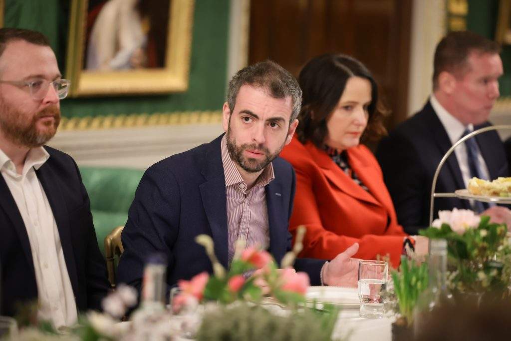  Several people seated along a dining table in an ornate room with green walls, floral centrepieces, and framed artwork.