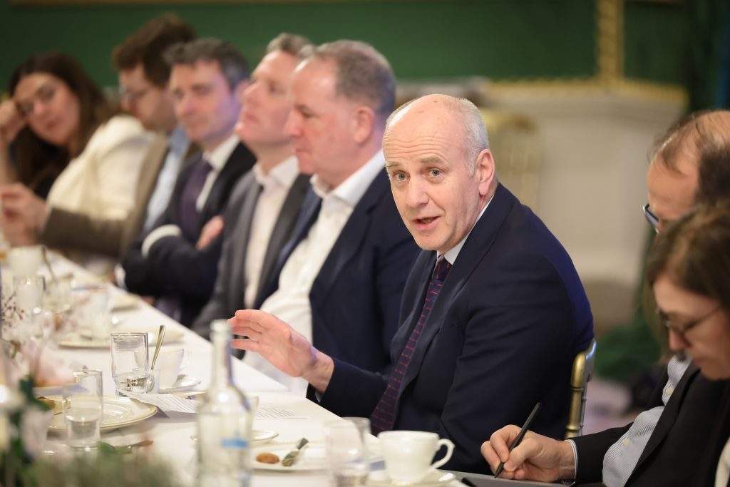 Several attendees seated at a long dining table, with one person gesturing while others listen. The room is decorated with elaborate green walls and paintings.