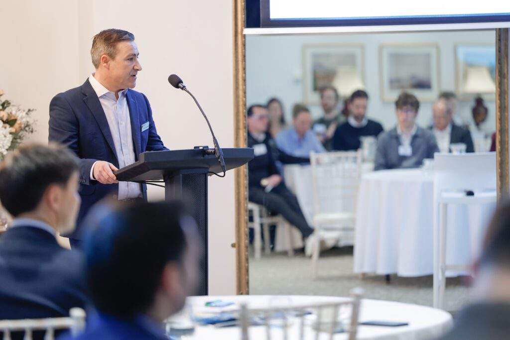 A speaker standing at a lectern addressing attendees, with a large mirror behind showing the audience seated at tables.