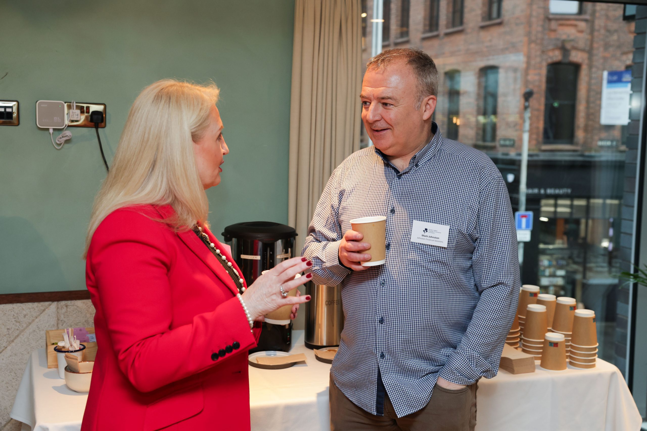 Two people standing beside a refreshment table, holding coffee cups and talking. A window and stacked paper cups are visible behind them.