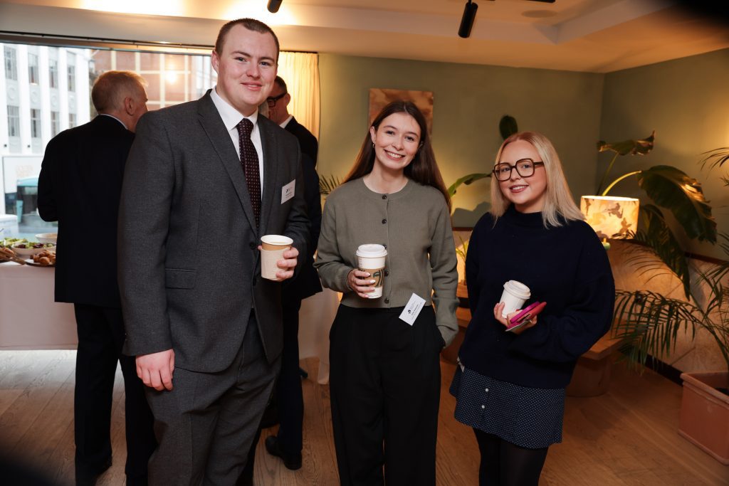 Three attendees standing together holding coffee cups, with a refreshments table and large window in the background.