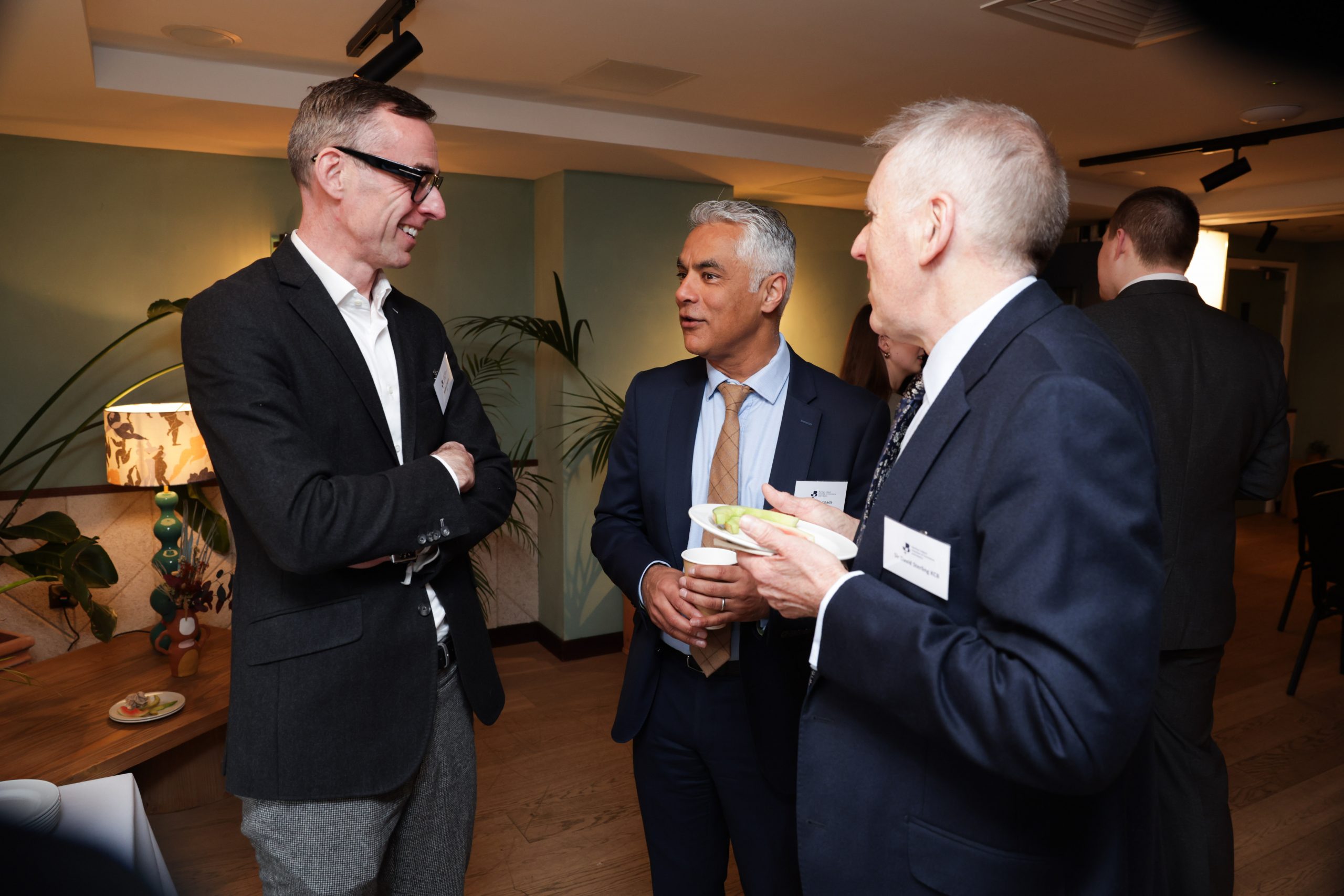 Three attendees engaged in conversation in a reception space, with one person holding a plate of food.