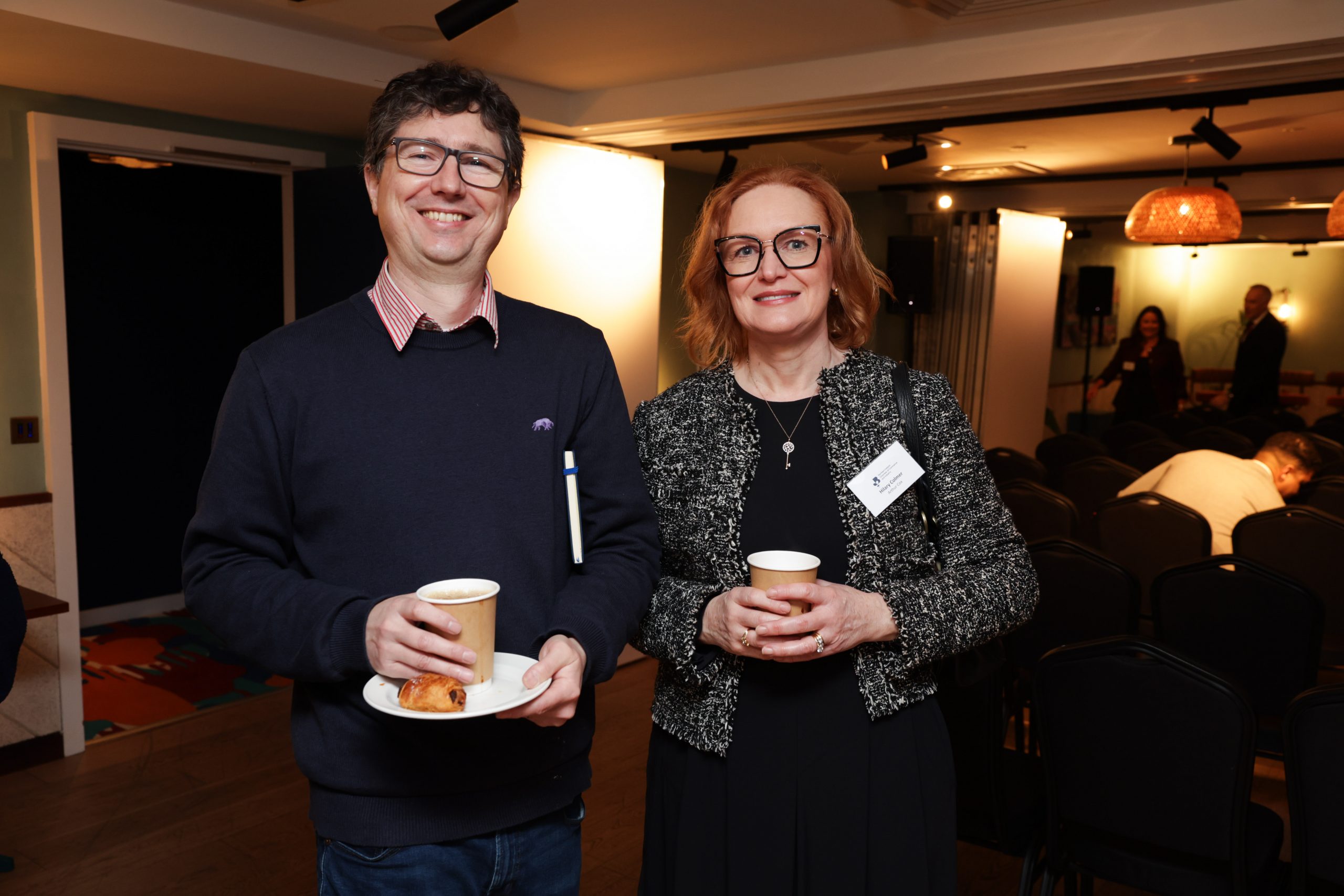 Two attendees standing side by side holding coffee cups, with rows of chairs and event lighting in the background.
