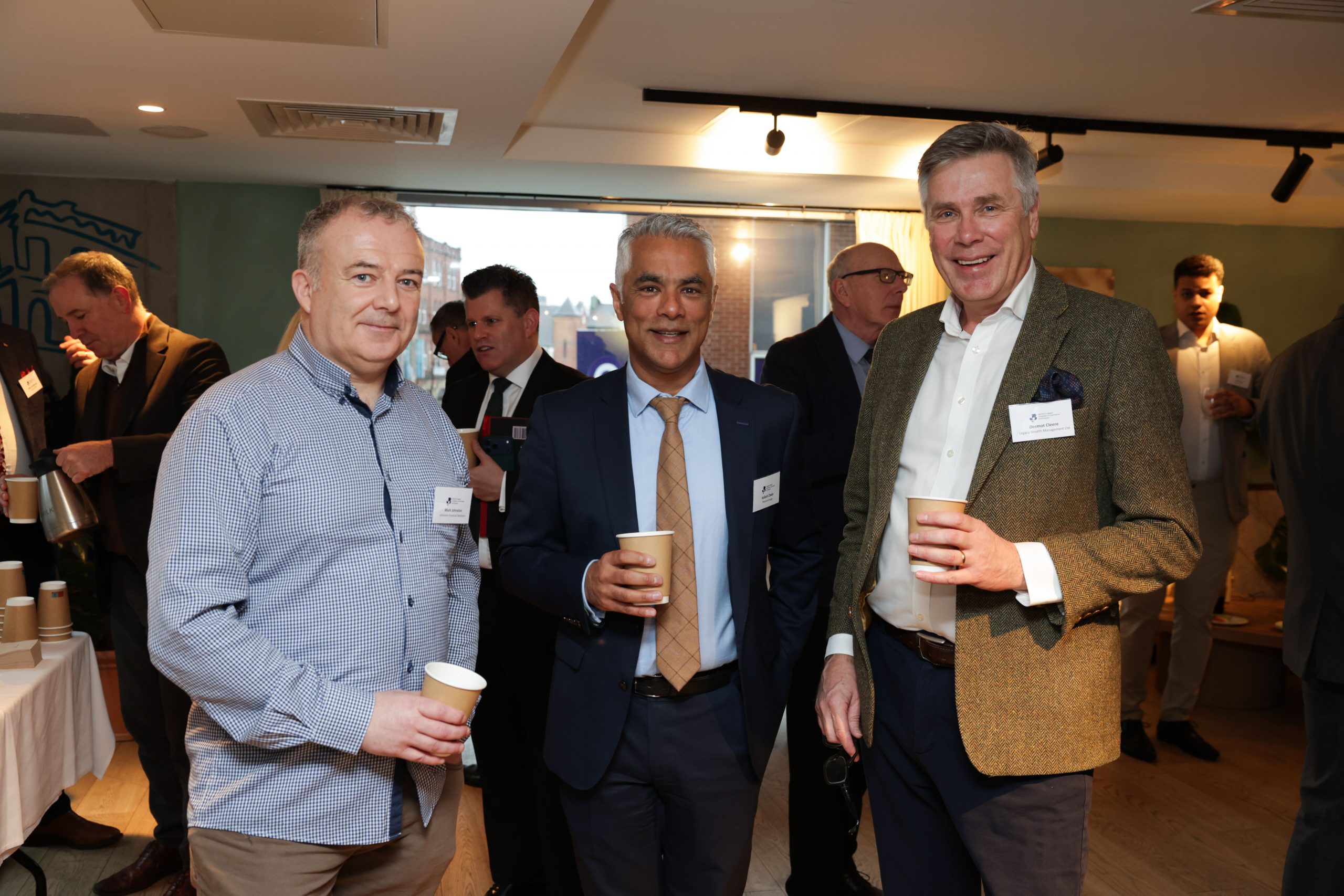 A group of three attendees standing together holding drinks during a networking session, with other guests visible behind them.