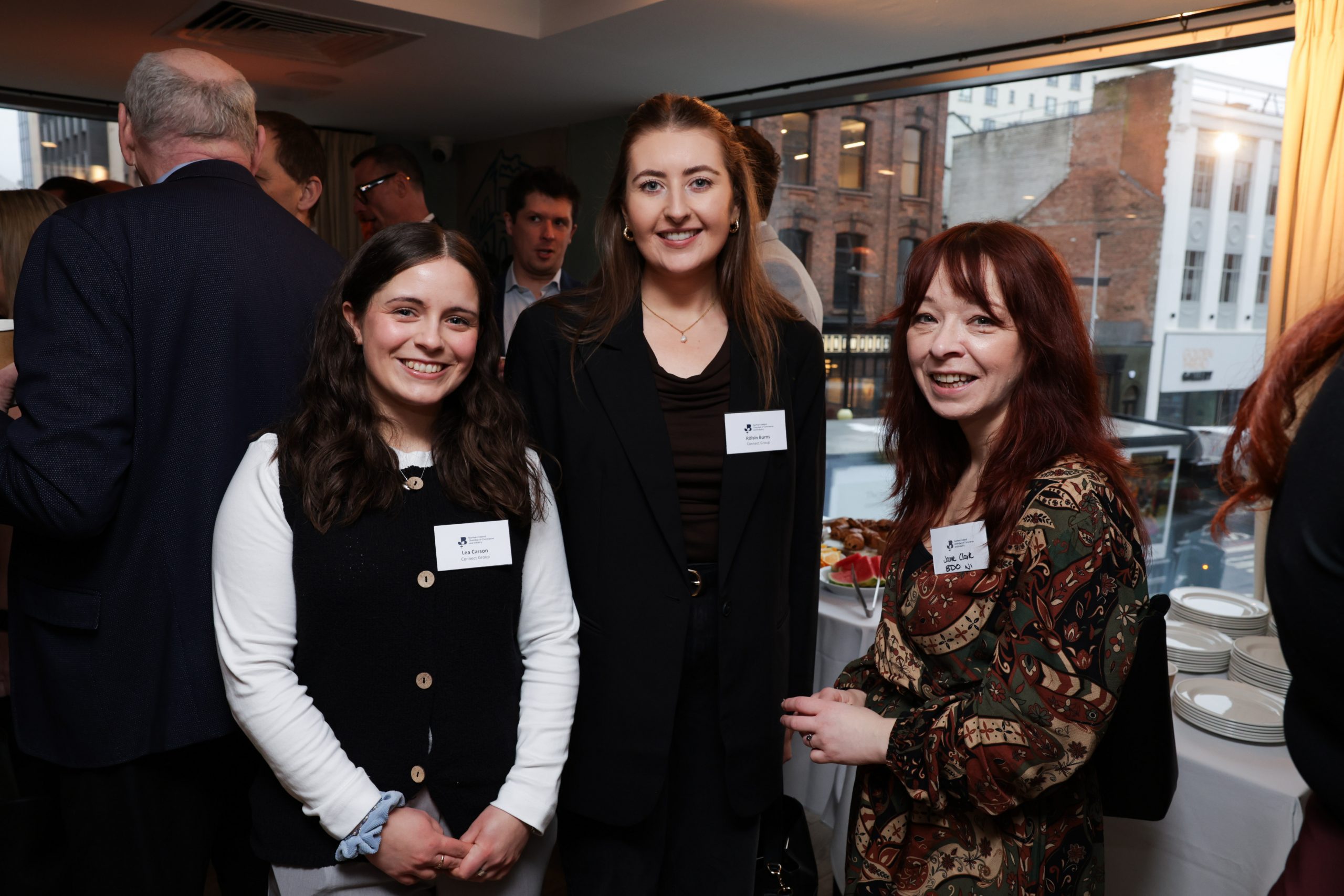 Three attendees standing together near a large window during a networking event, with a city street visible outside