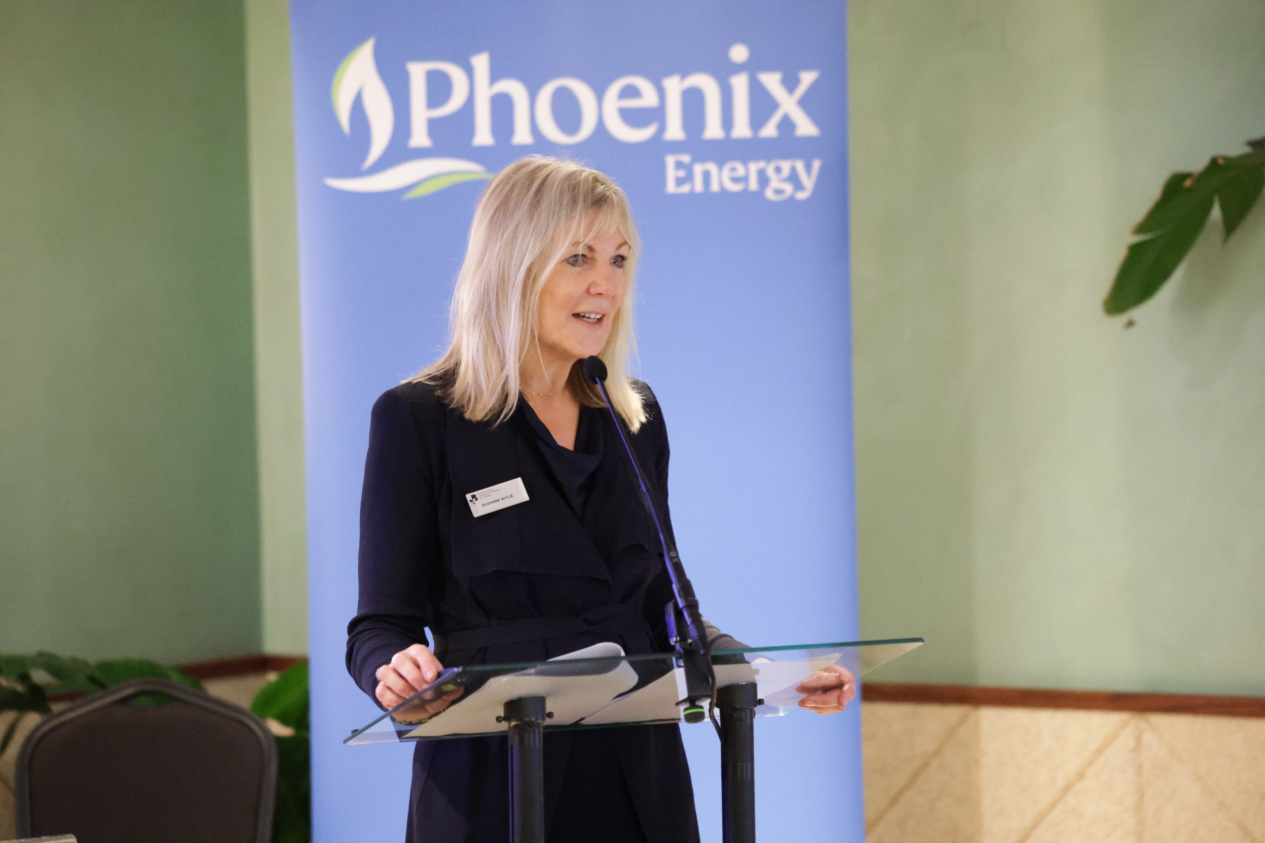 A speaker standing behind a glass lectern with a “Phoenix Energy” banner behind them, holding notes while presenting.