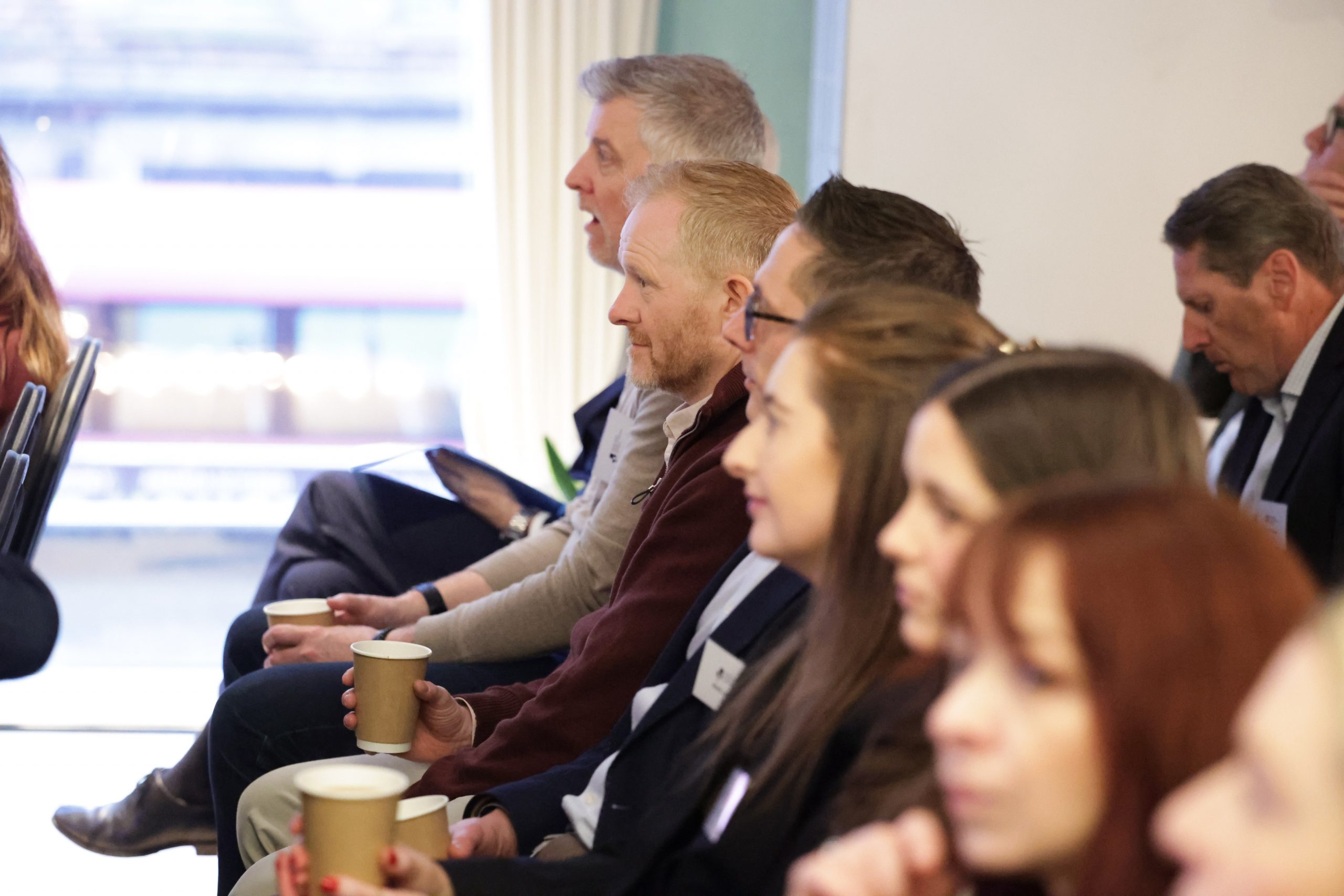 A row of attendees seated and holding coffee cups while watching an event.