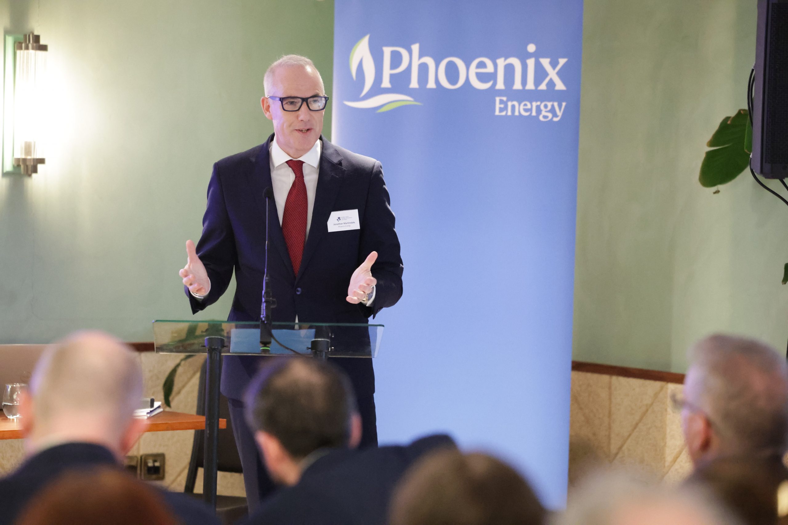 A speaker standing behind a lectern addressing an audience, with a “Phoenix Energy” banner behind them.
