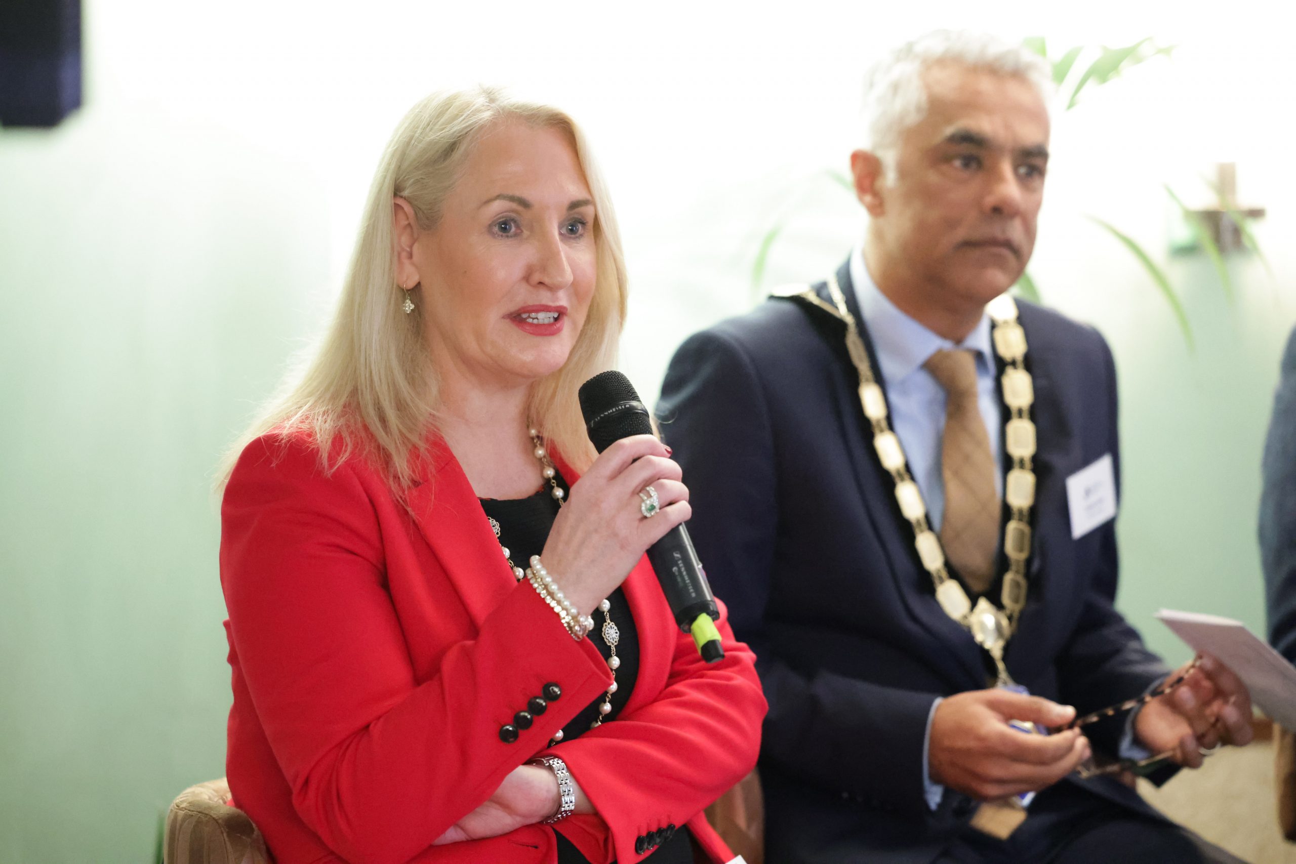 A panel discussion in progress, with two seated speakers holding microphones. One person is wearing a chain of office.
