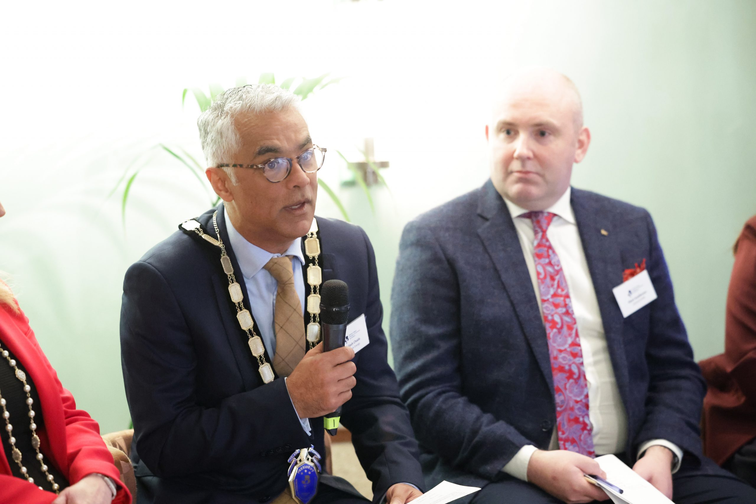 Two panel speakers seated next to each other. One speaker holds a microphone, and another wears a chain of office.