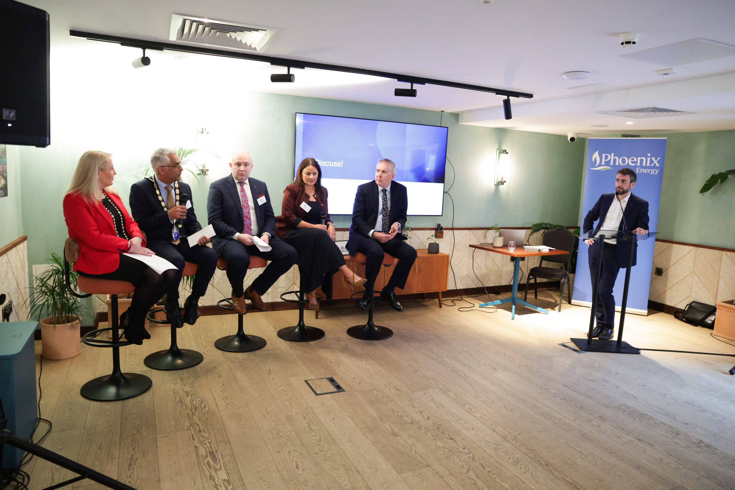 A panel of five speakers seated on high stools at the front of a room, with a presenter standing at a lectern and a screen displaying a presentation behind them.
