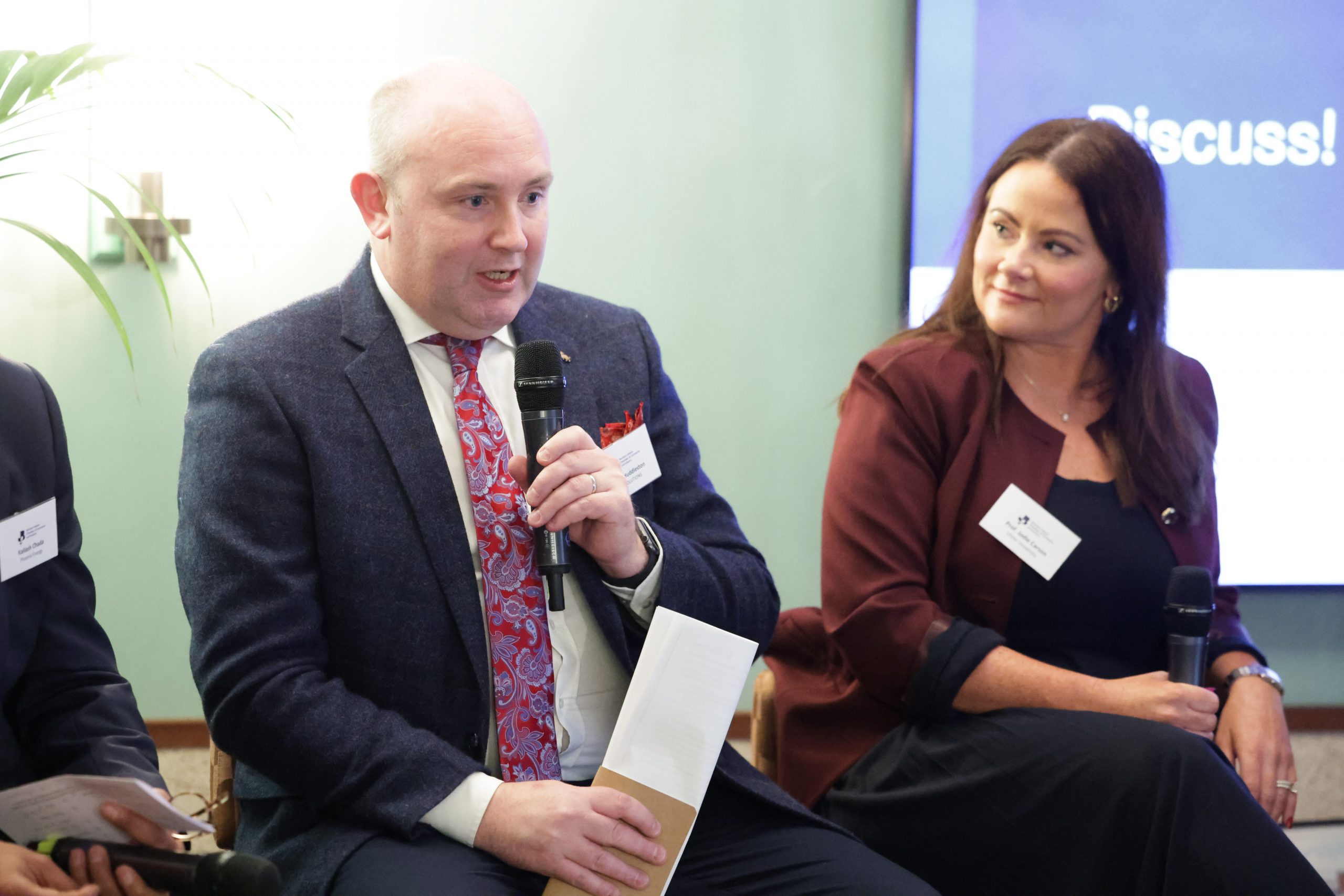 A close-up of two panel speakers seated next to each other, each holding a microphone and notes.