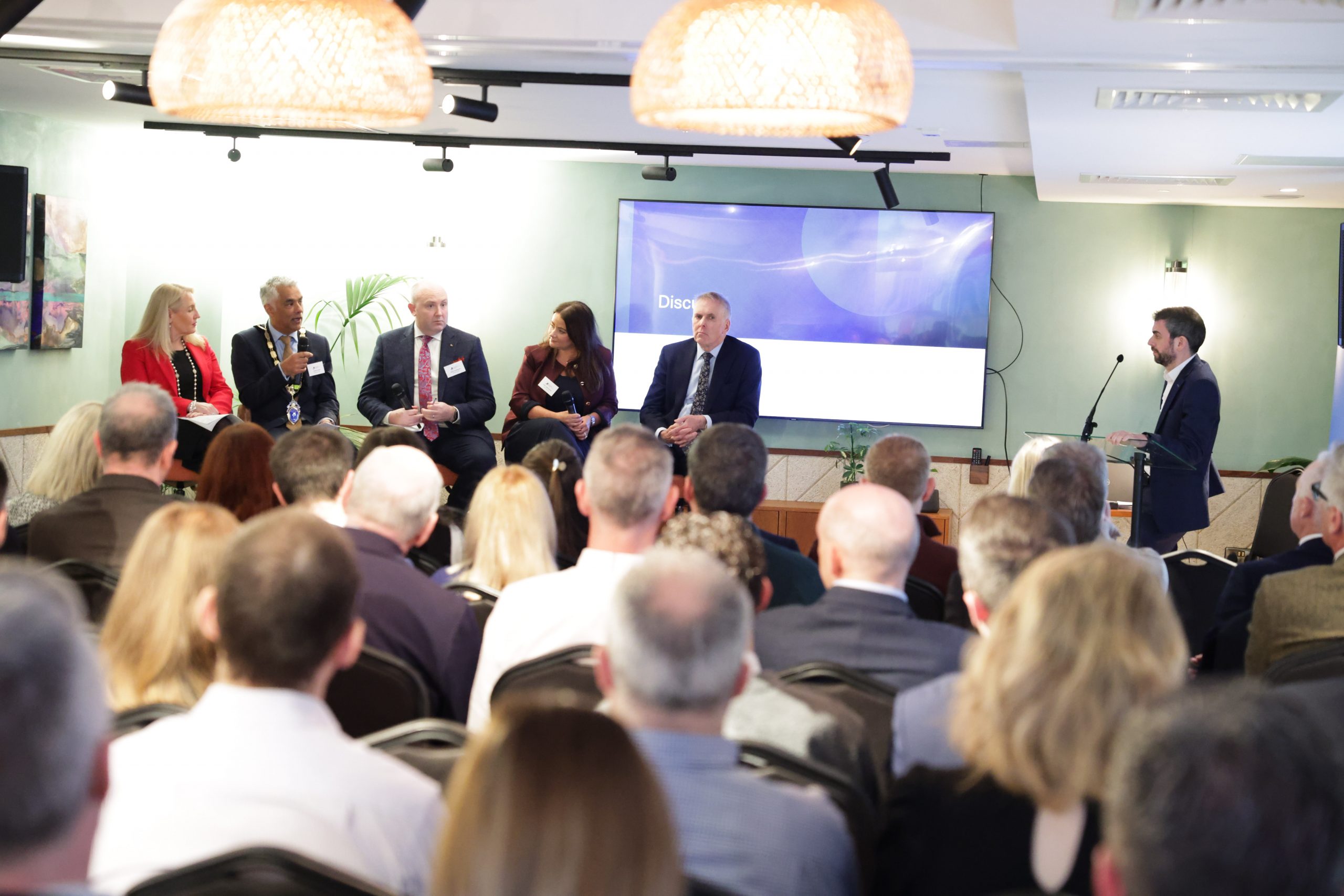 A room filled with attendees facing a panel discussion at the front. Five speakers are seated beneath a presentation screen while an additional presenter stands at the side.