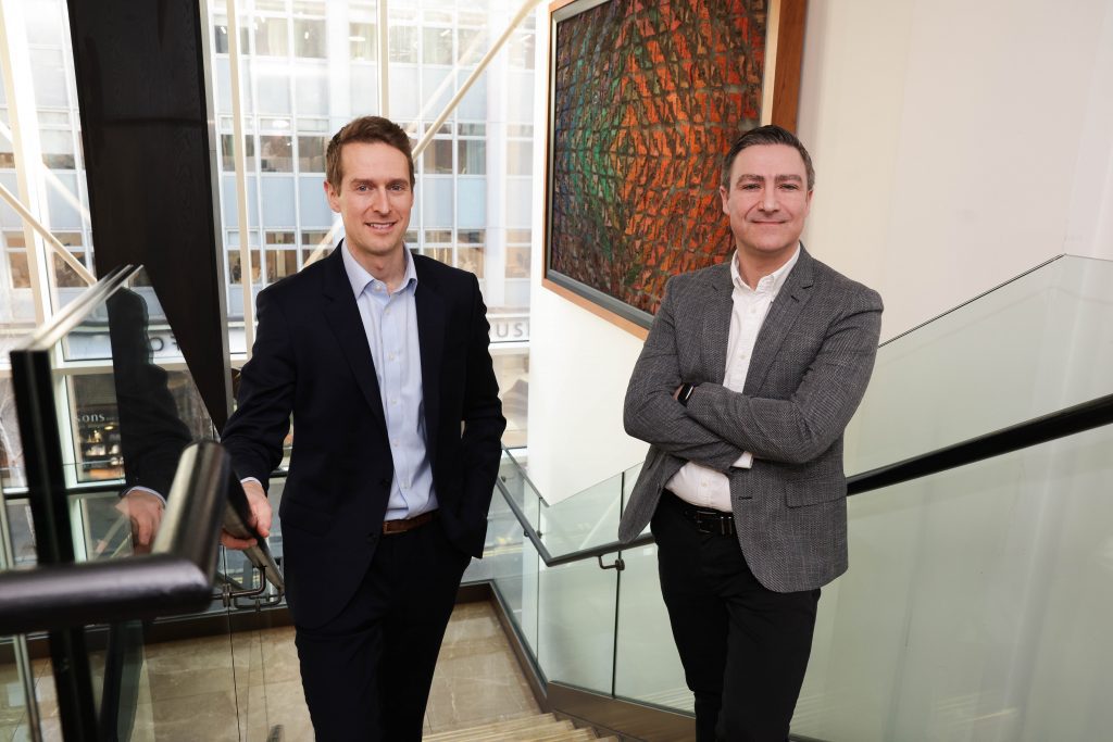 Two professionally dressed individuals stand on a staircase inside a modern building with glass panels and artwork on the wall.