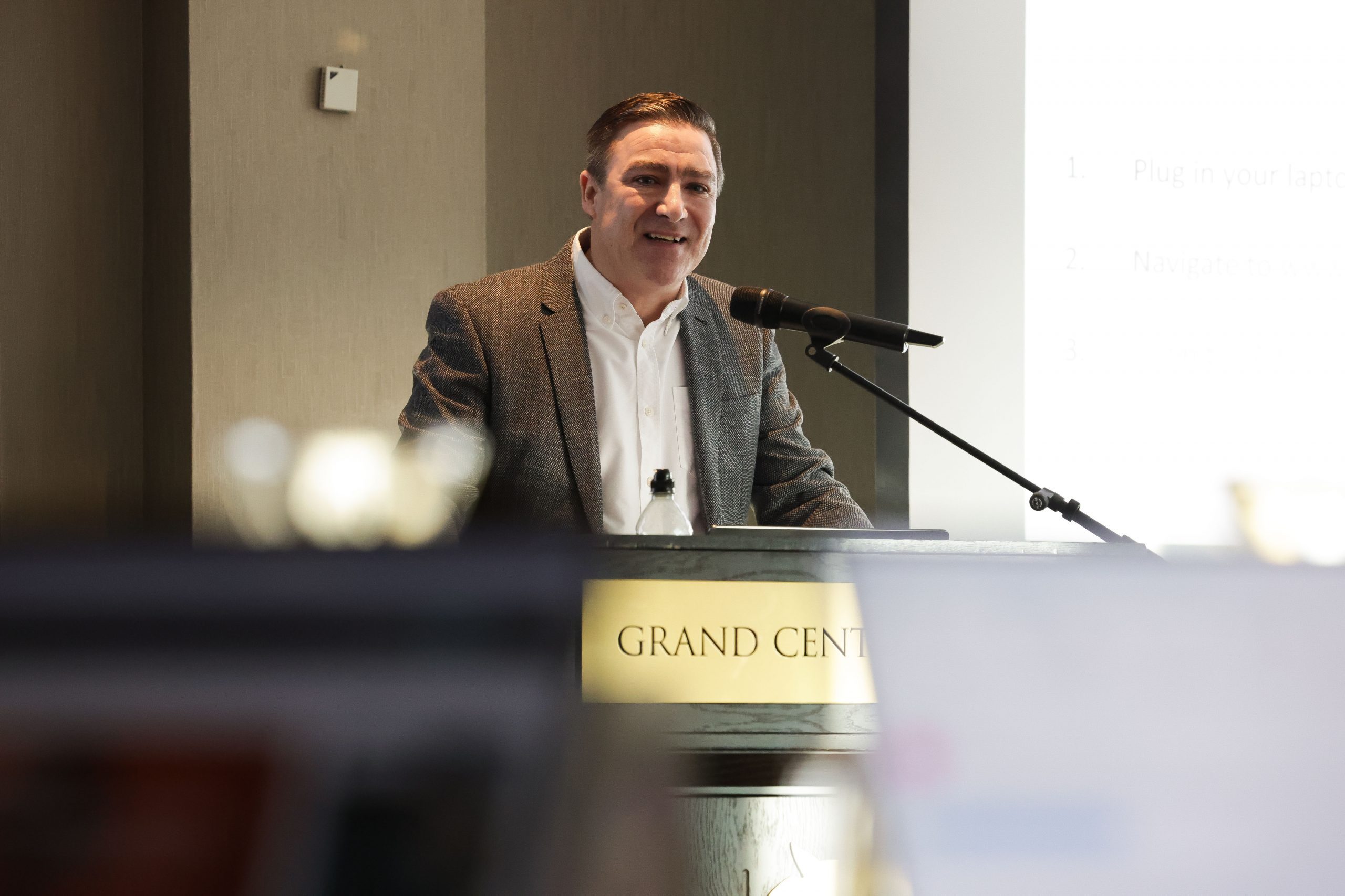 A speaker stands at a lectern labelled “Grand Central,” delivering a presentation with projected slides in the background.