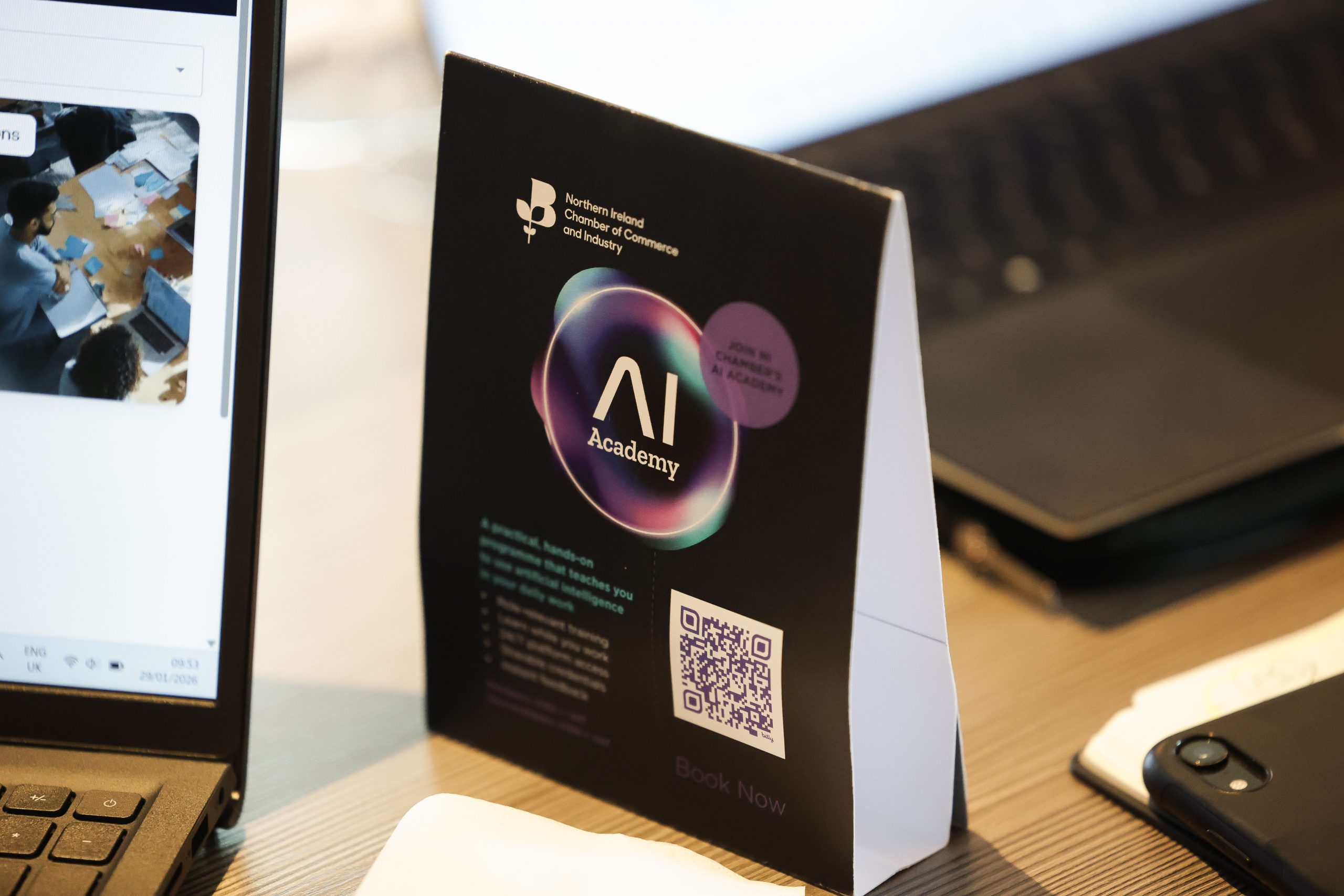 A table display card promoting the “AI Academy” sits beside open laptops and other digital devices.