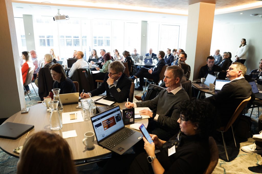 A large conference room filled with attendees sitting at round tables, all engaged with laptops and presentation materials.