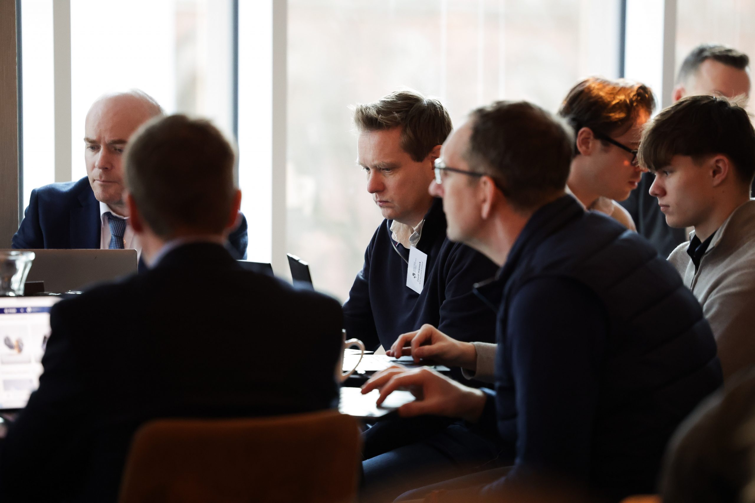 A group of participants sit around a table engaged in conversation and using laptops at a professional workshop.