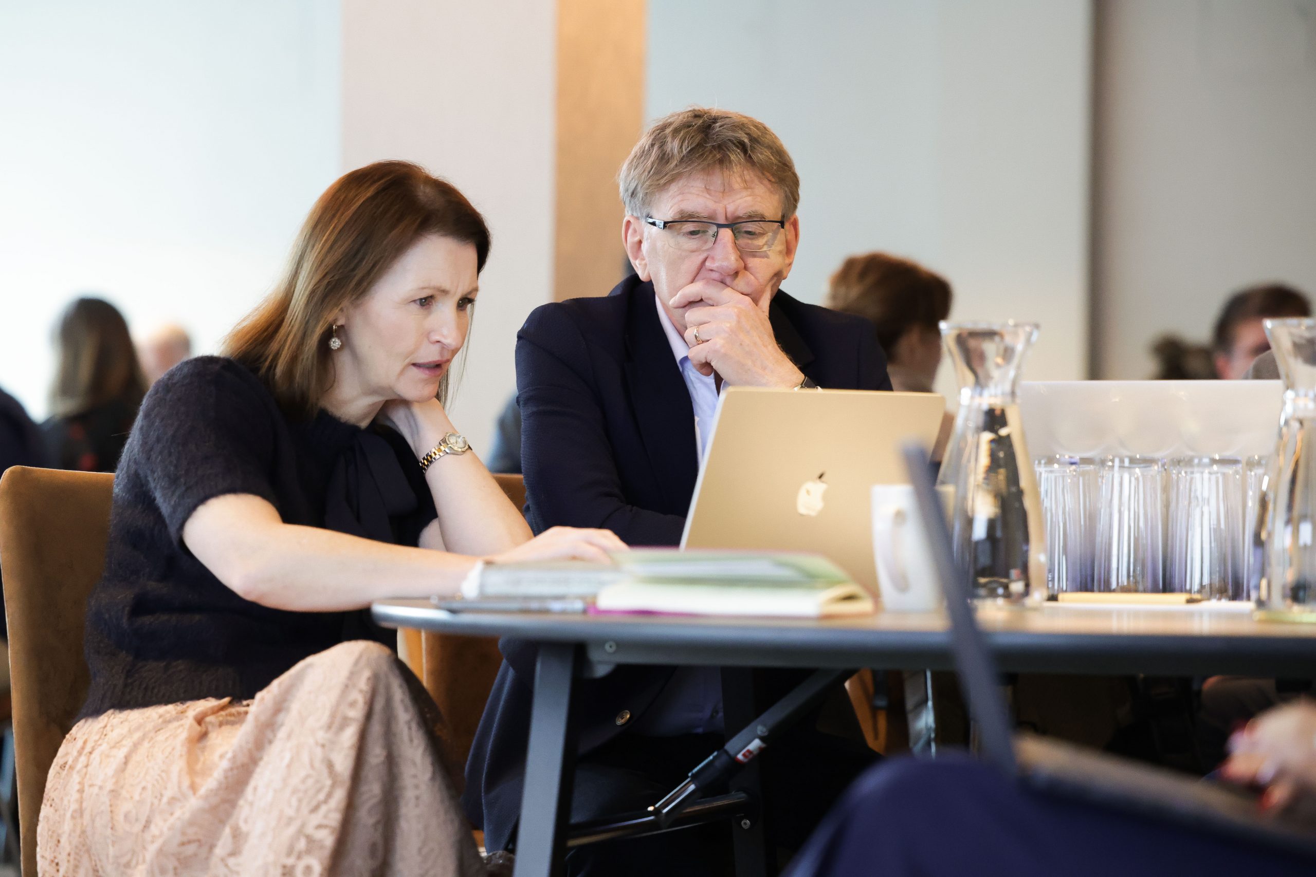 Two colleagues sit at a round table working on a laptop and documents during a business event.