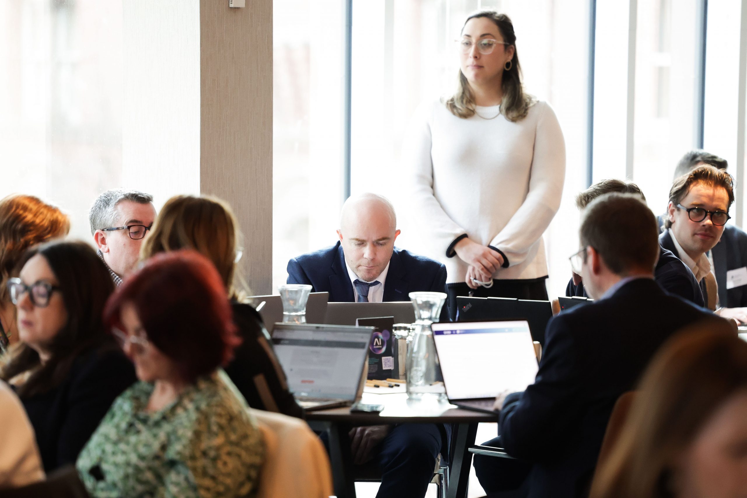A woman standing beside a seated group addresses the room, while participants listen and work at round tables near large windows.