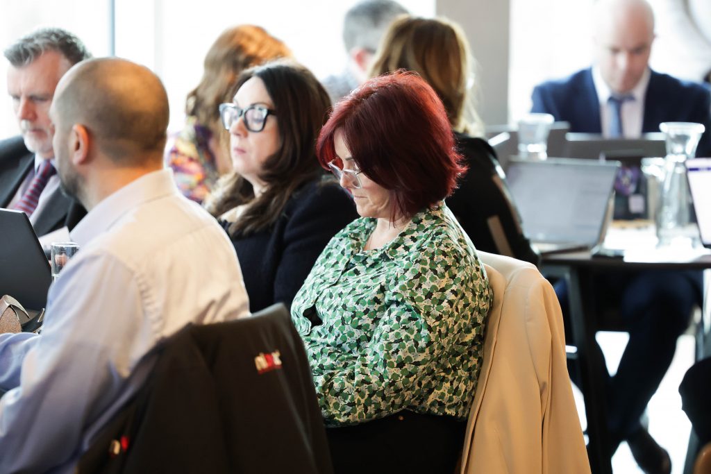  A woman in a patterned green top sits among a group of attendees during a busy workshop session.