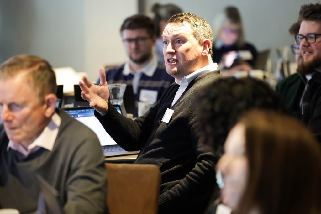 A person seated in a conference room gestures with one hand while others around them listen and work on laptops