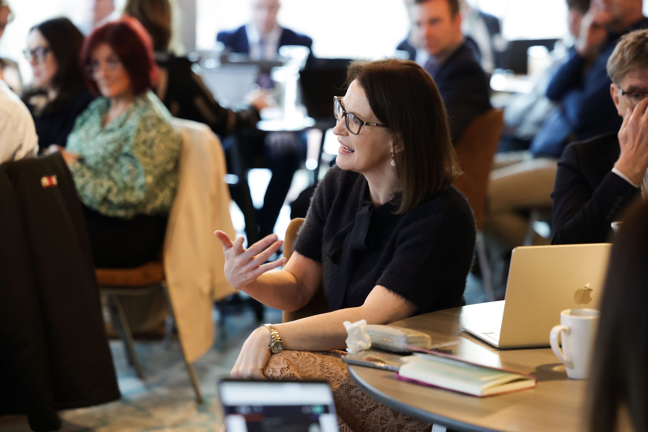 A woman seated at a conference gestures as she speaks, with notebooks, laptops and attendees around her.