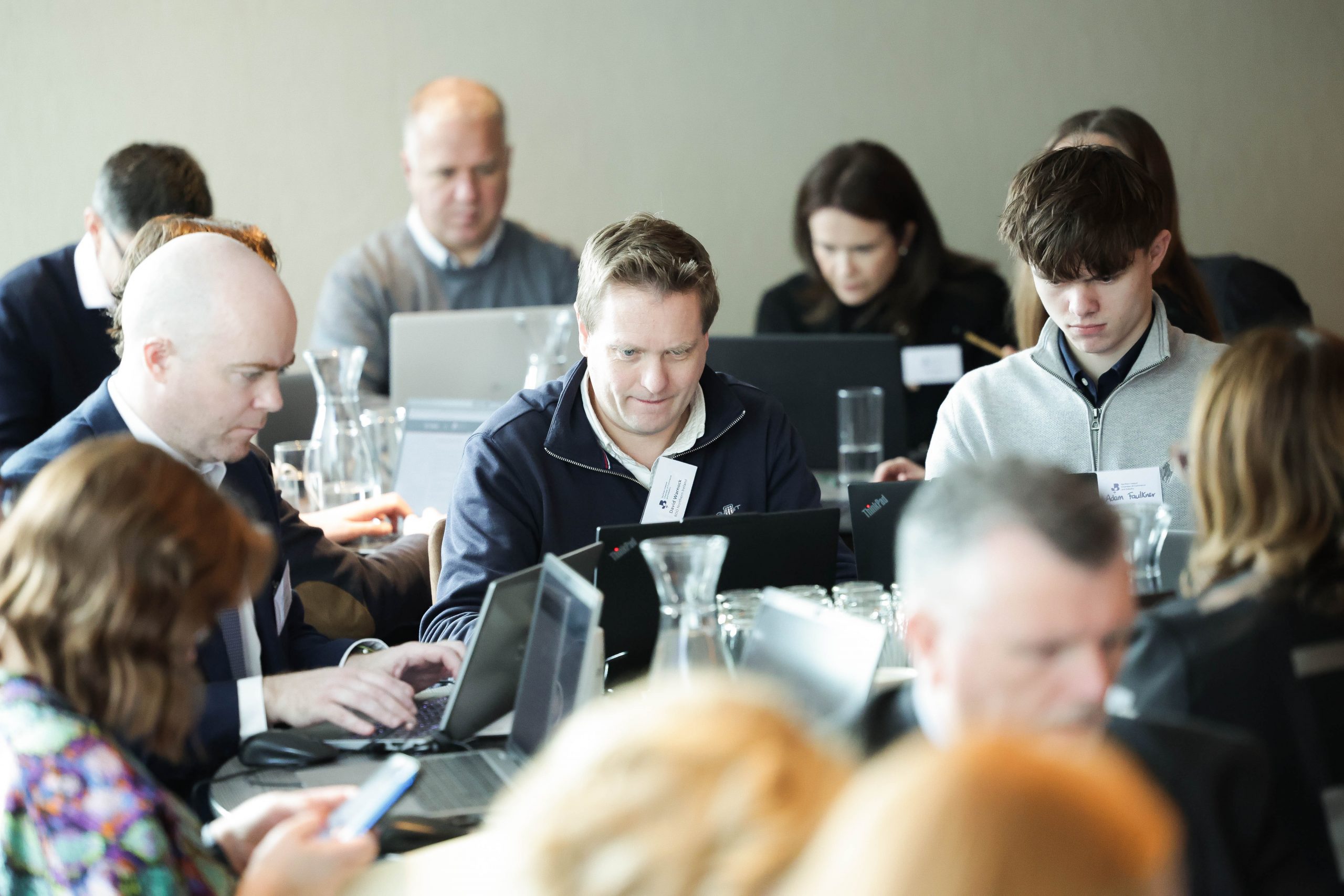 A group of attendees sit closely together at tables, focused on laptops and notes during a workshop-style event.