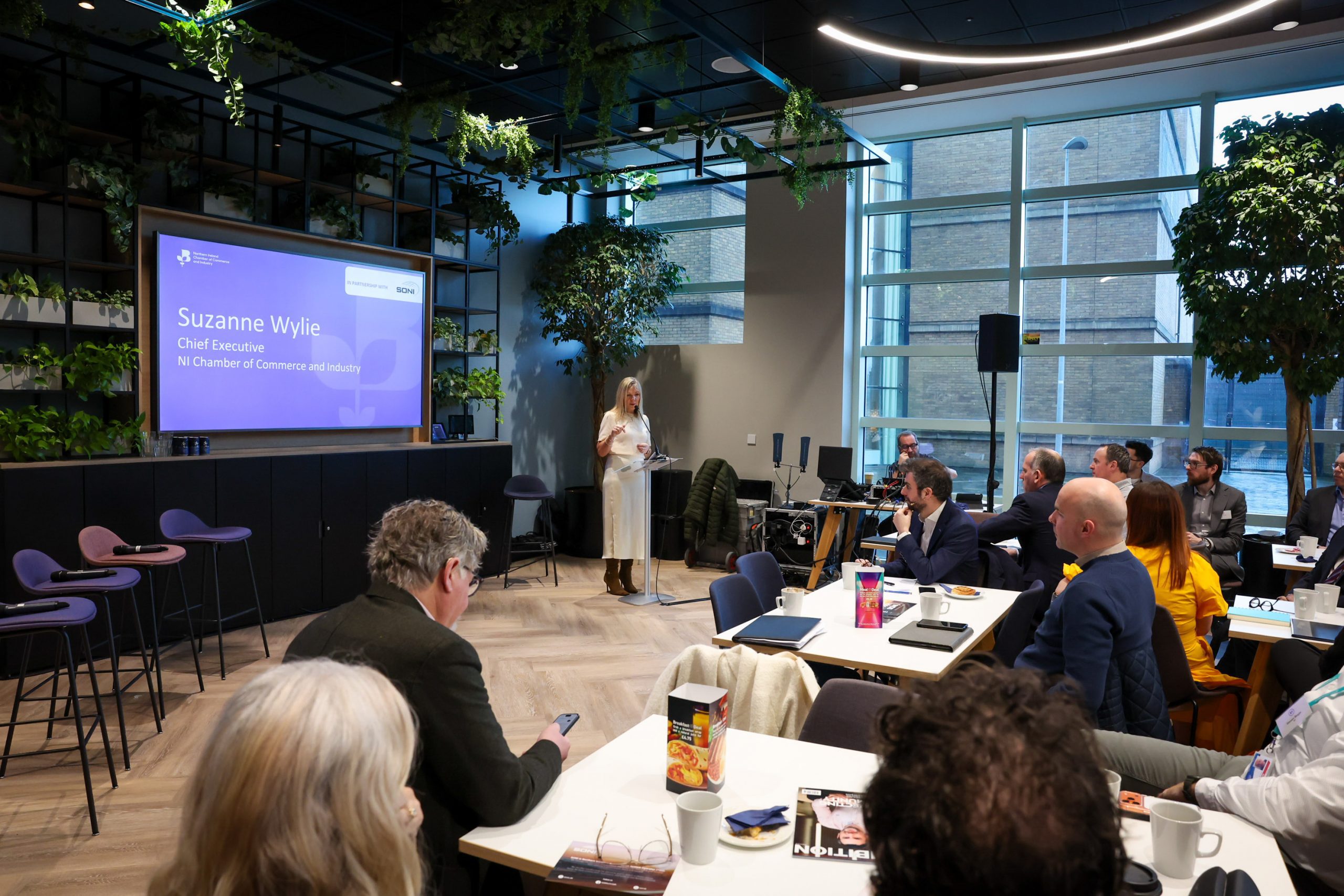 A woman presents on stage beside a large screen showing her name and title.