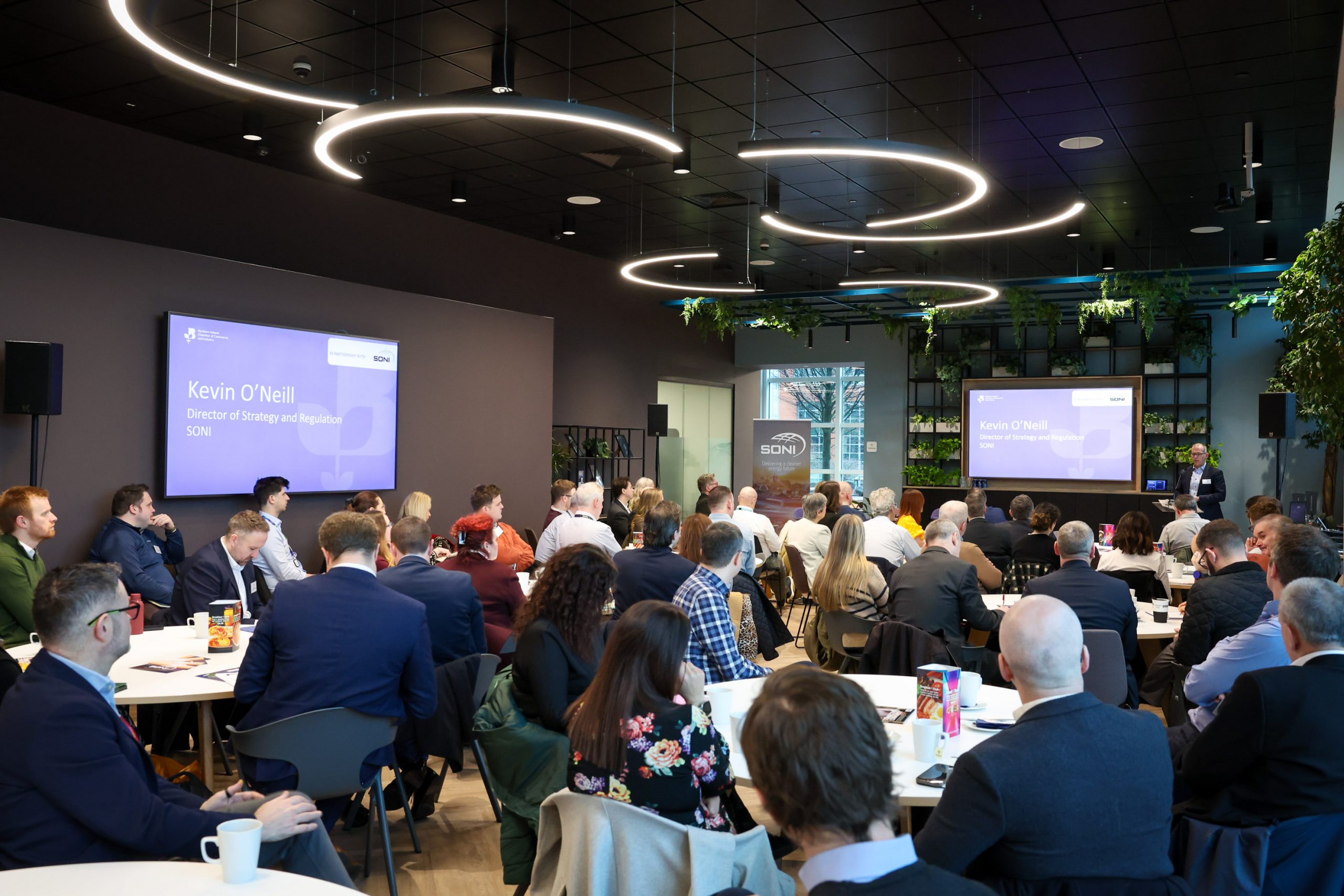A large audience watches a presentation displayed on two screens in a conference room.