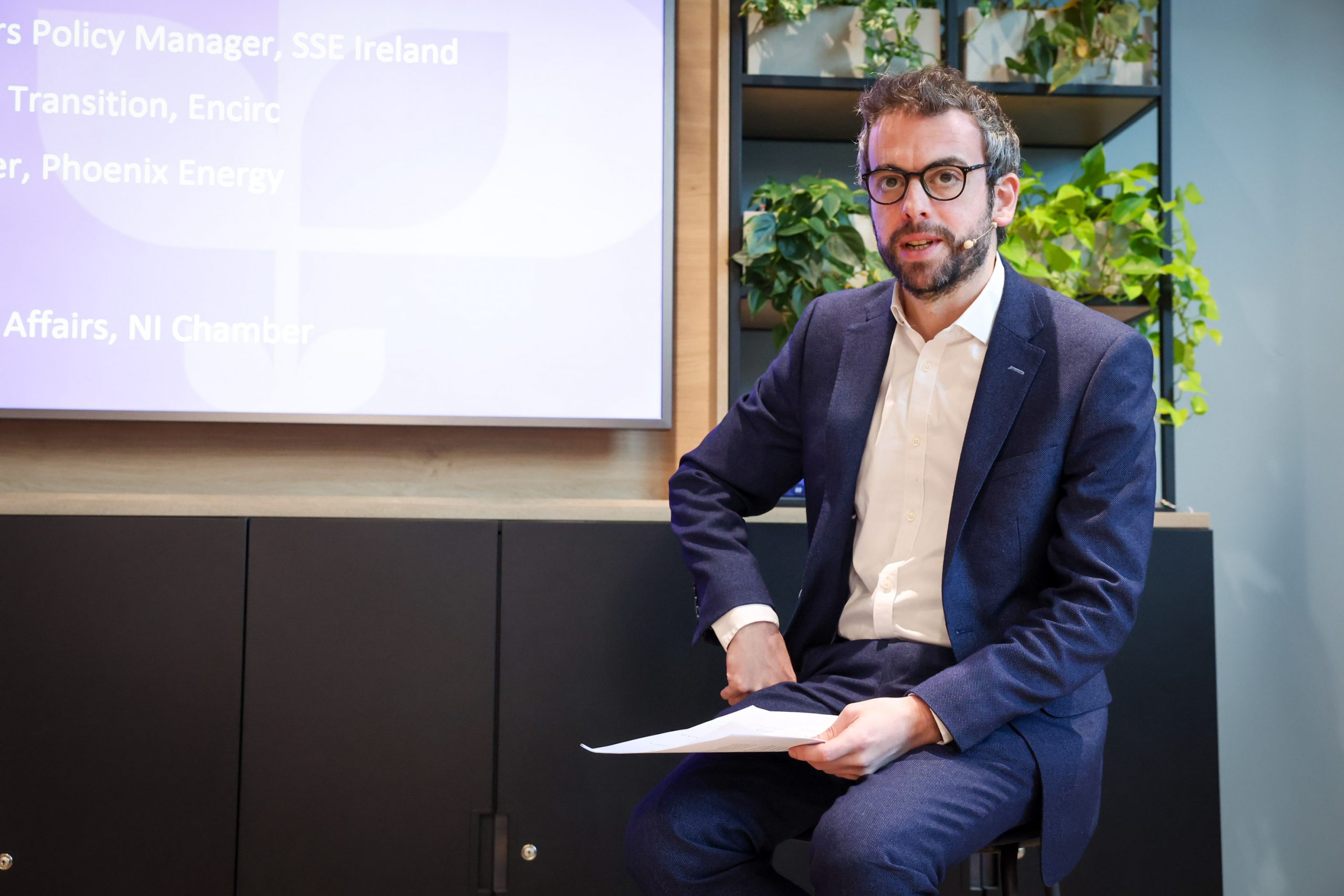 A person in a suit sits on a stool holding papers beside a presentation screen