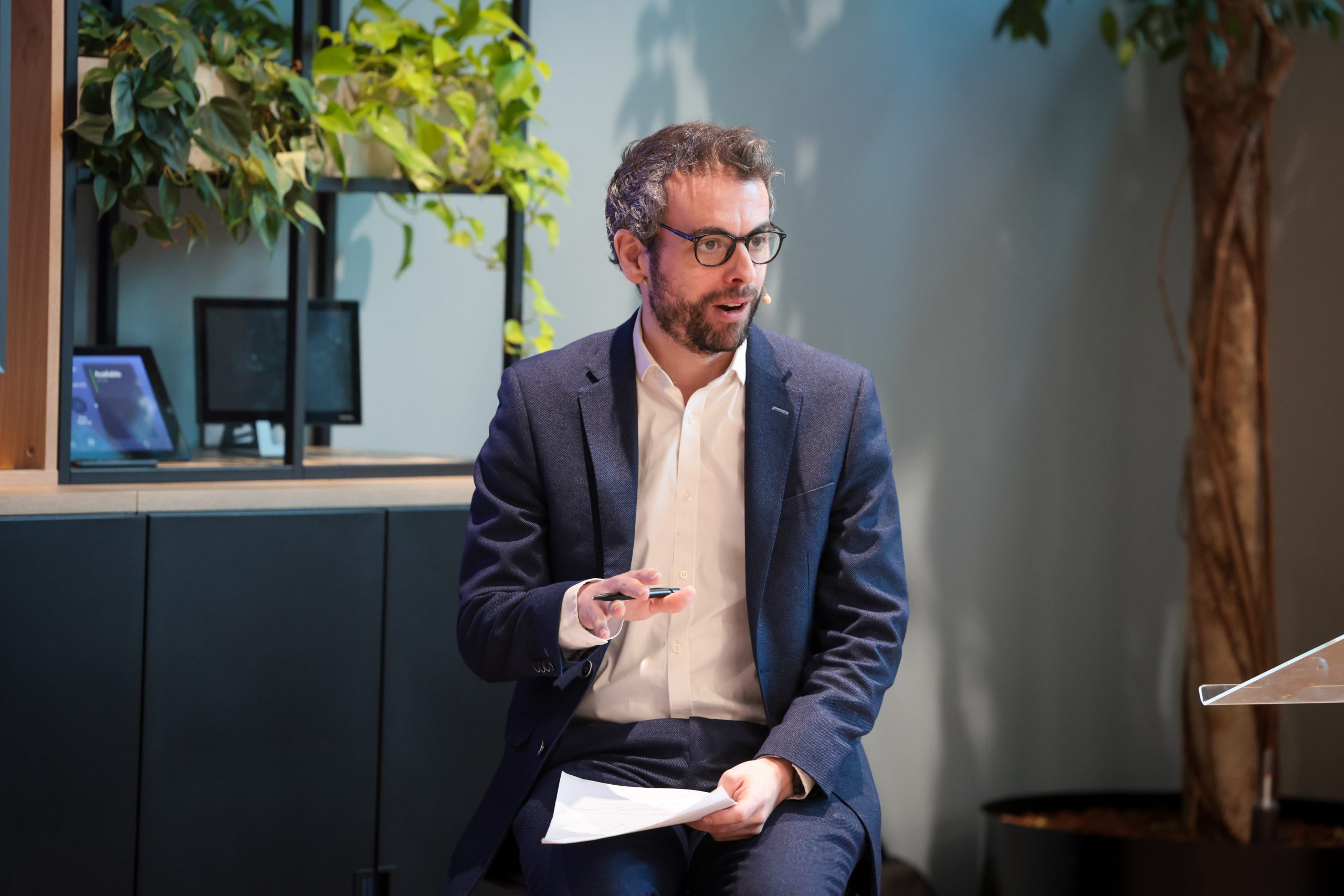 A person in a suit sits holding papers and a remote during a talk.