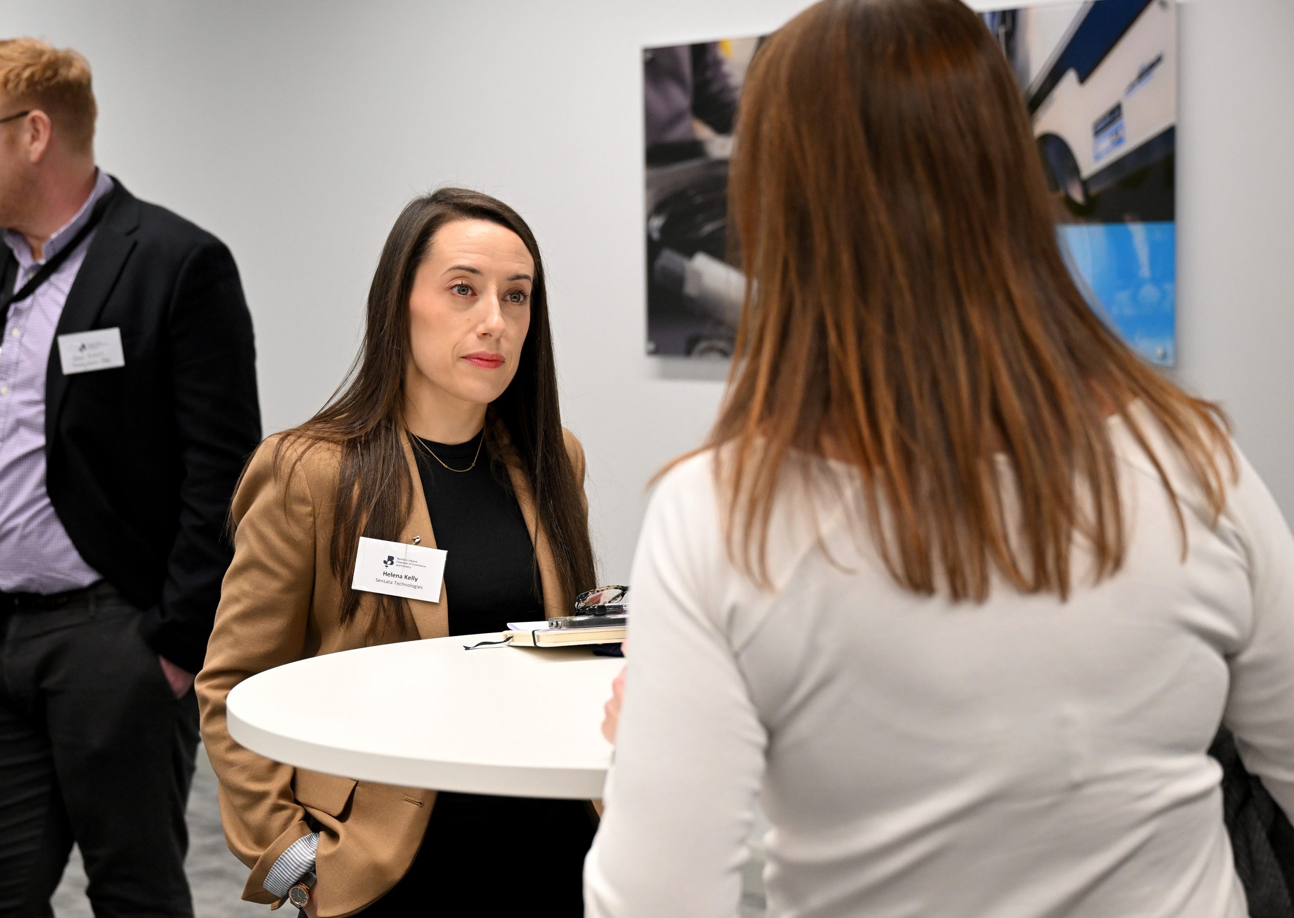 Two people chat at a standing table during a networking event, with another attendee in the background.