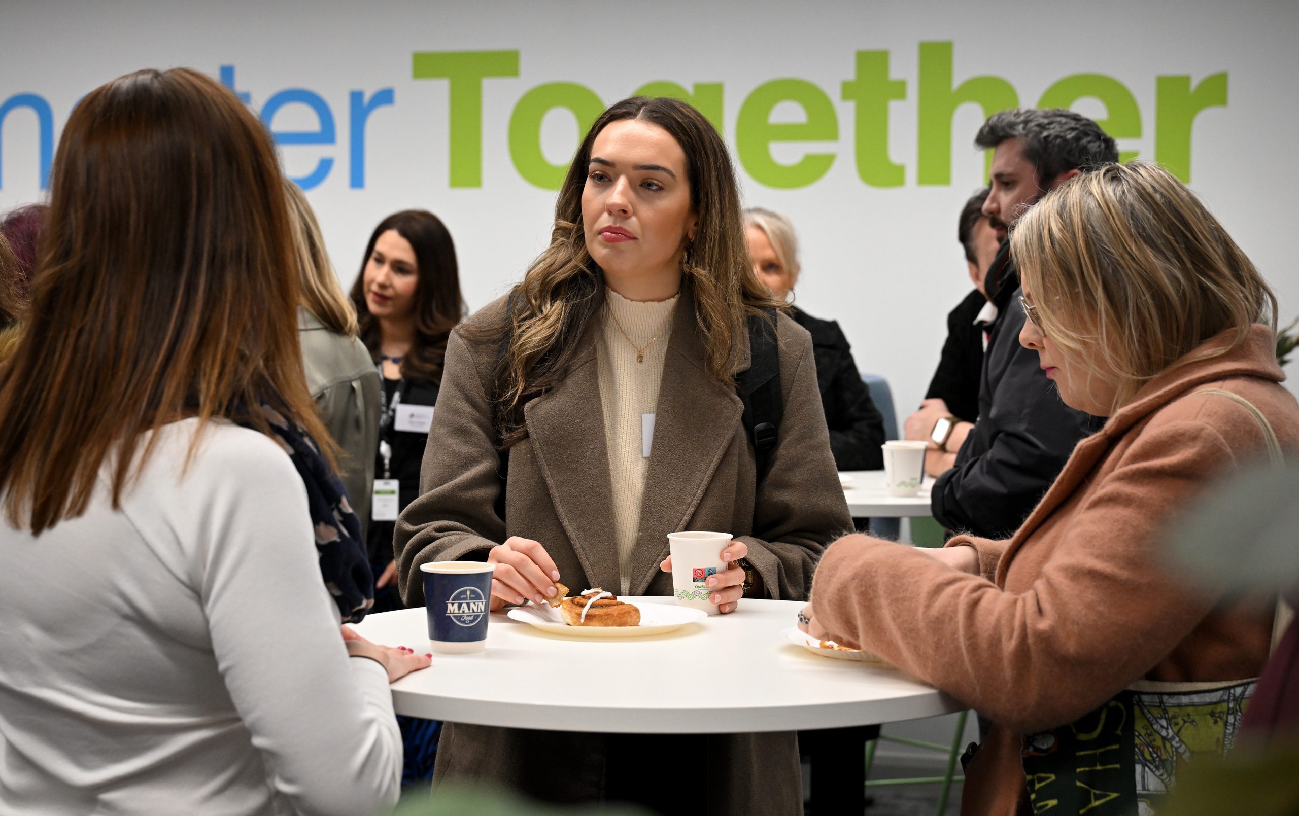 A group of attendees stand around a tall table enjoying coffee and pastries, with a large “Together” wall graphic behind them.