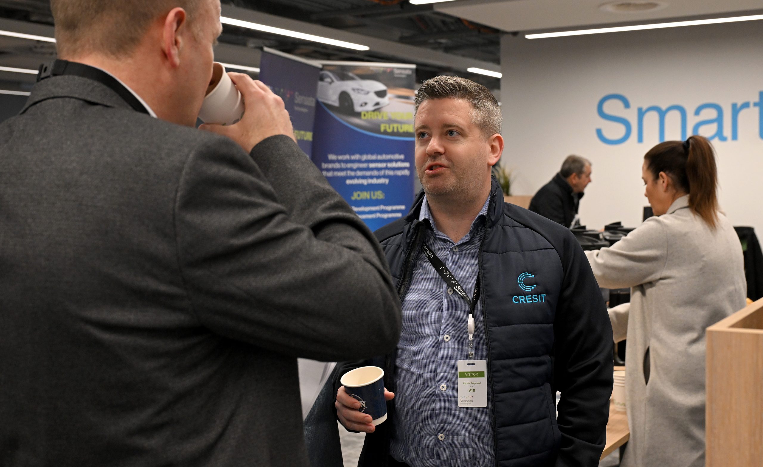 Two attendees talk while holding coffee cups near event banners promoting an automotive technology company.