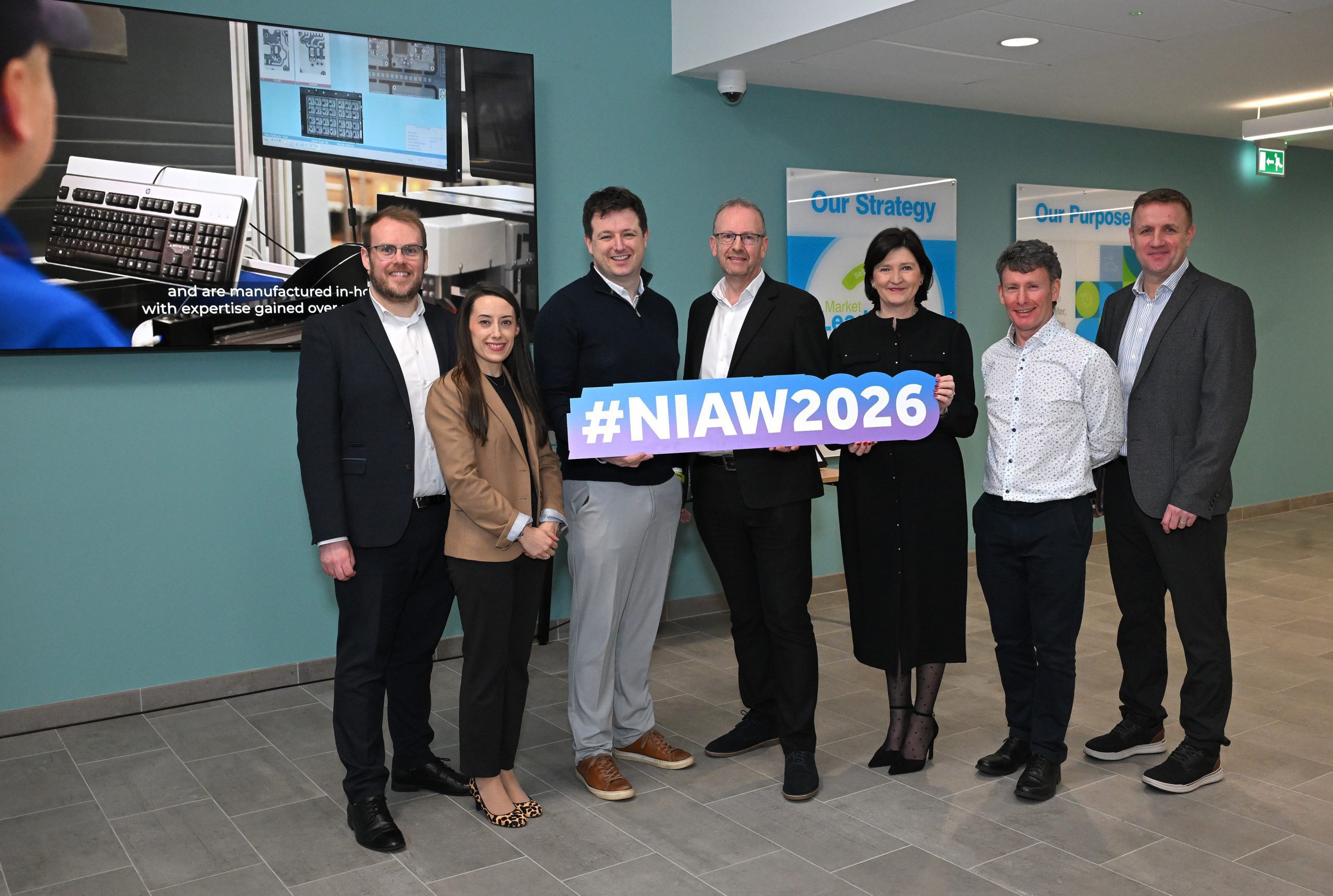 A group photo of several attendees holding a large “#NIAW2026” sign inside an office lobby.