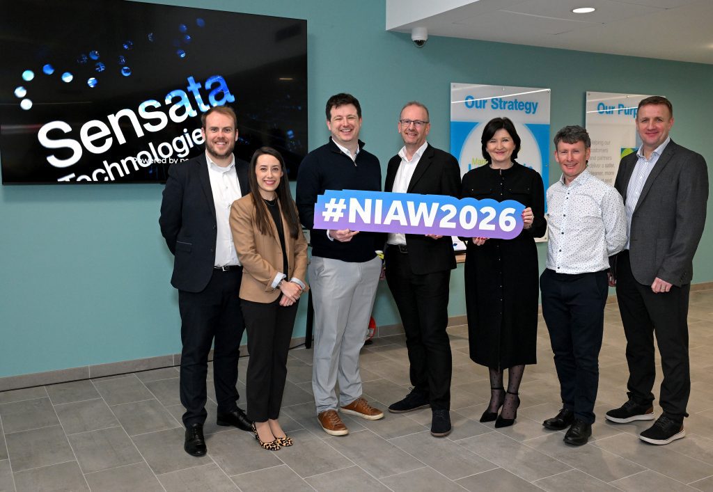  A group of attendees pose for a photo holding a “#NIAW2026” sign in front of a wall displaying a company logo.