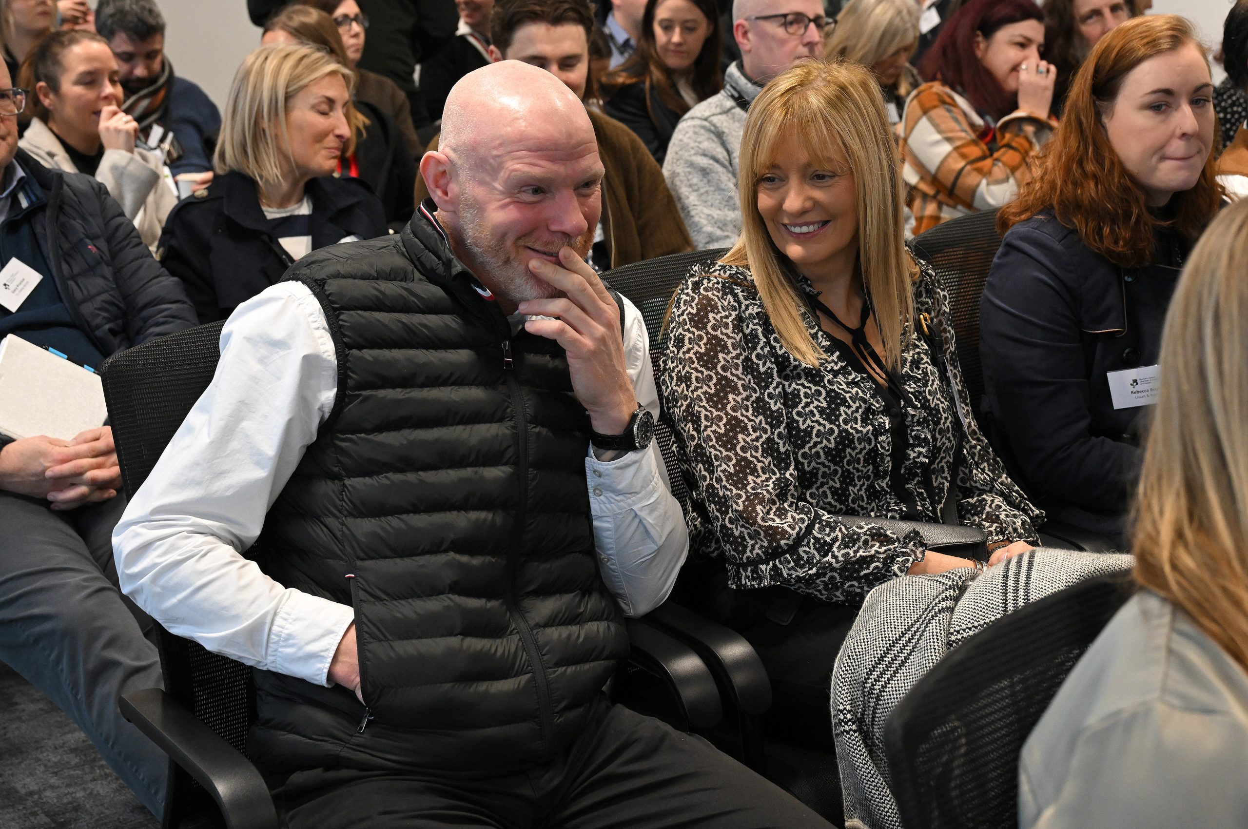 Two attendees sit listening closely during an event, one holding a hand to their face thoughtfully.